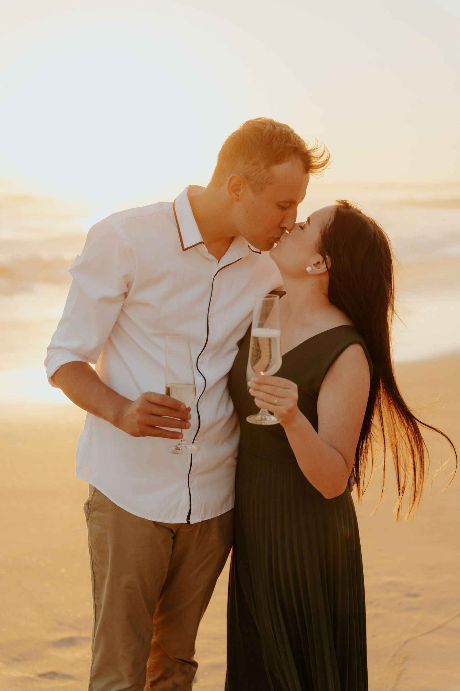 Couple kissing on Mosselbay beach at sunset holding champagne glasses, woman in green dress, man in white shirt.