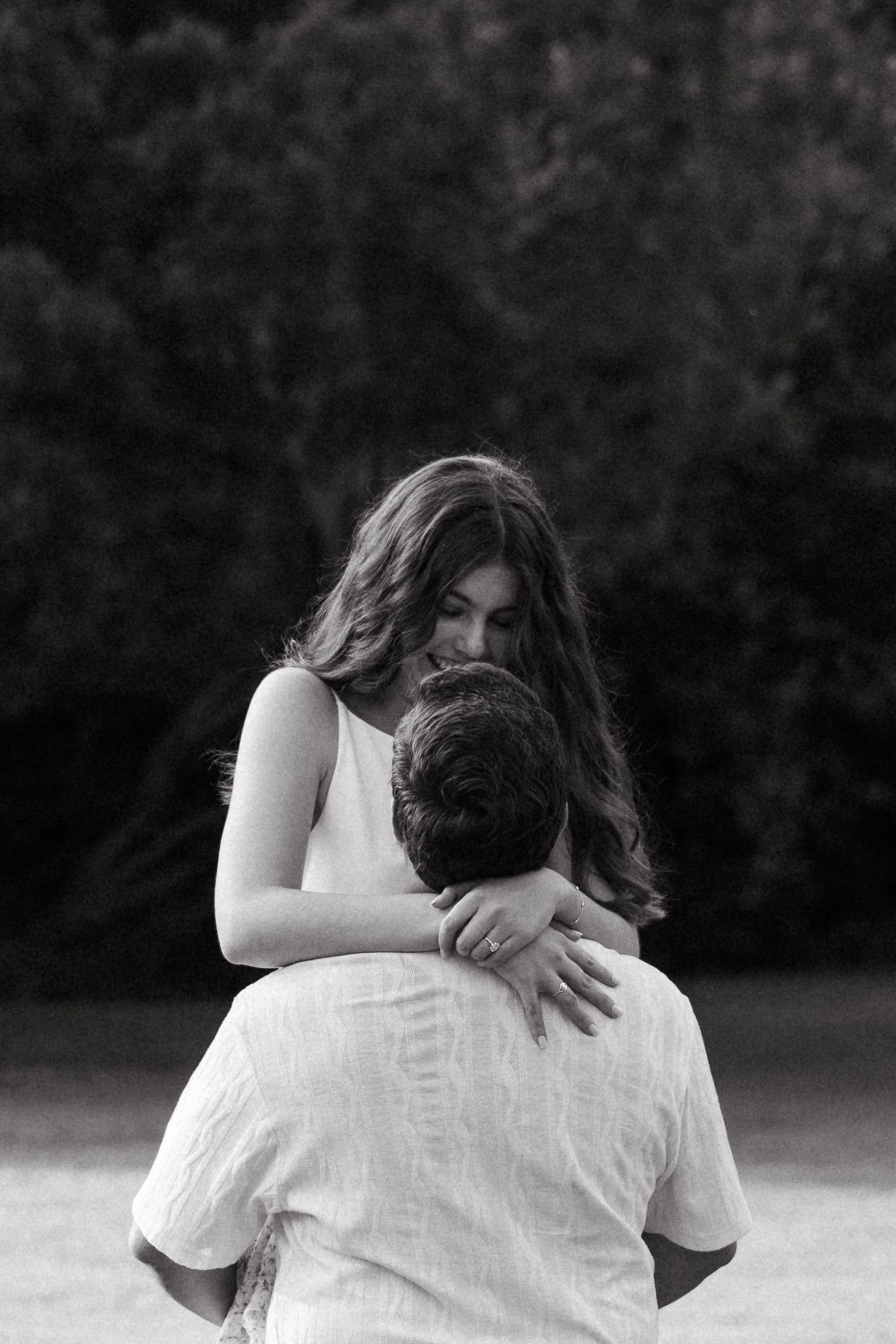 Black and white photo of a couple embracing outdoors, woman wrapping arms around mans neck with trees in background.