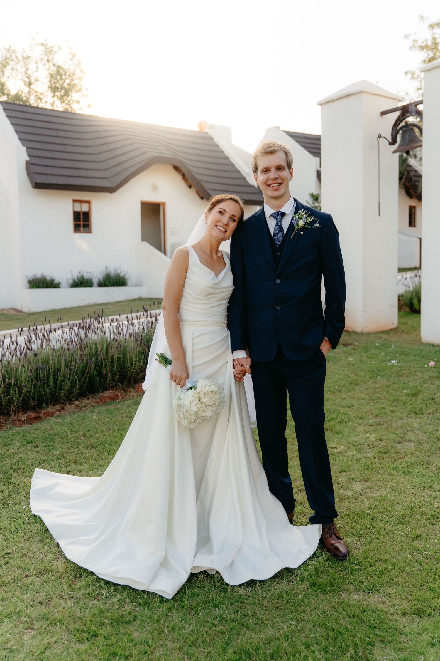 A couple poses playfully together on a grassy lawn in front of a white building during their wedding day.