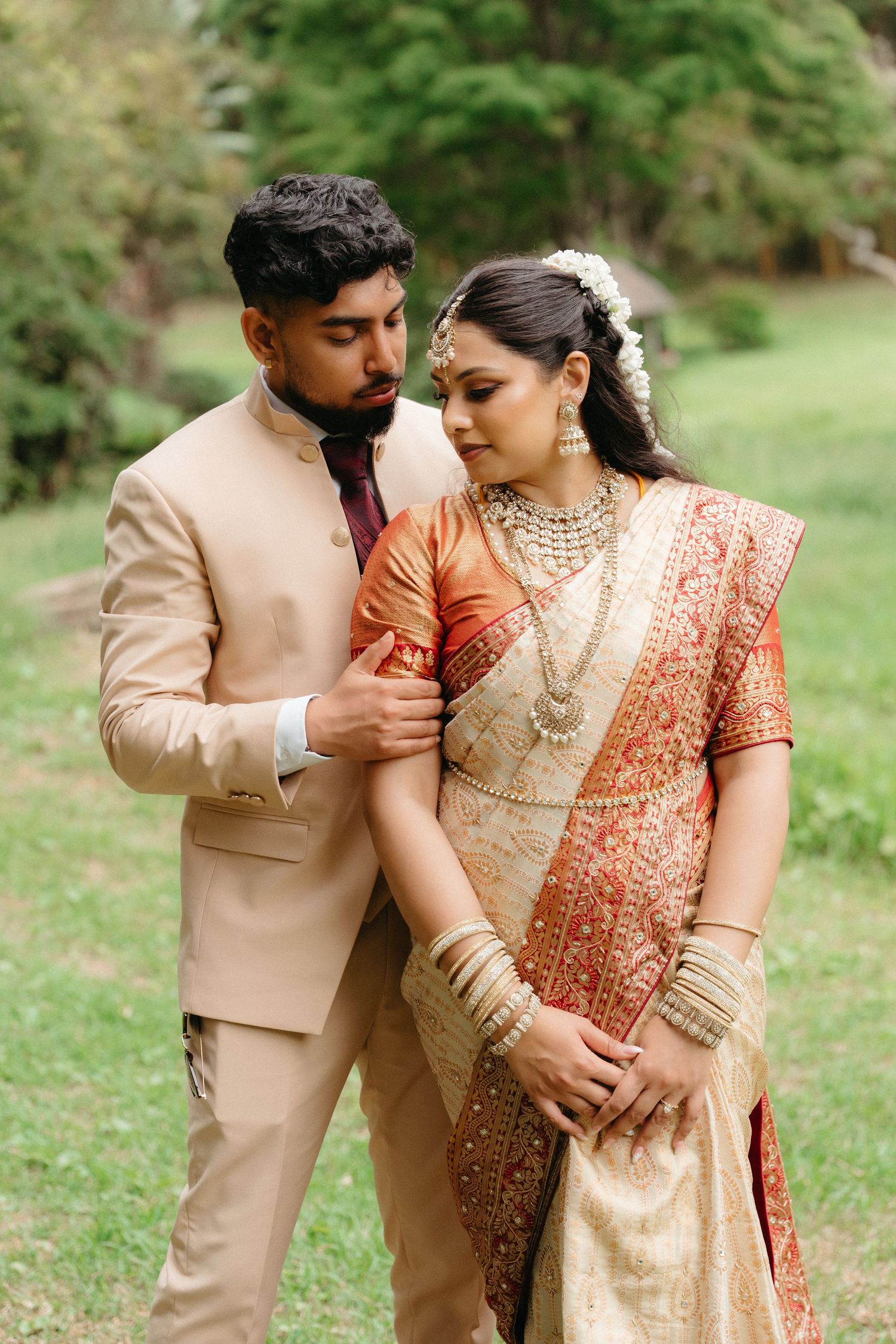 A couple in traditional South Asian wedding attire embrace outdoors, wearing beige and coral formal clothing with gold jewelry.