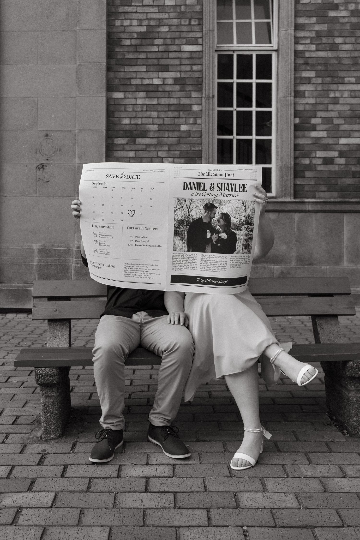 A couple sits on brick steps sharing a newspaper, with their faces hidden behind the open pages in black and white.