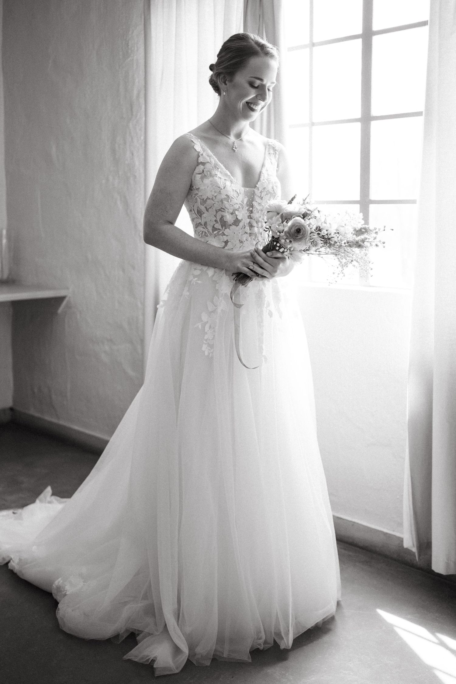 Black and white portrait by window showing elegant lace wedding gown and delicate bouquet.