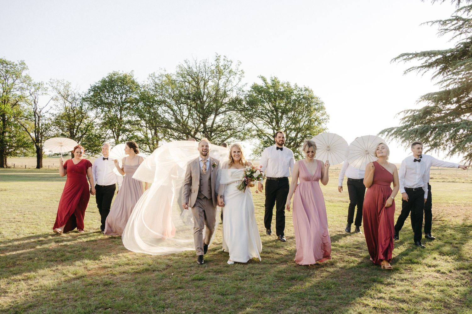 Bridal party poses joyfully on a sunlit lawn with flowing dresses catching the breeze during a golden hour photo session.