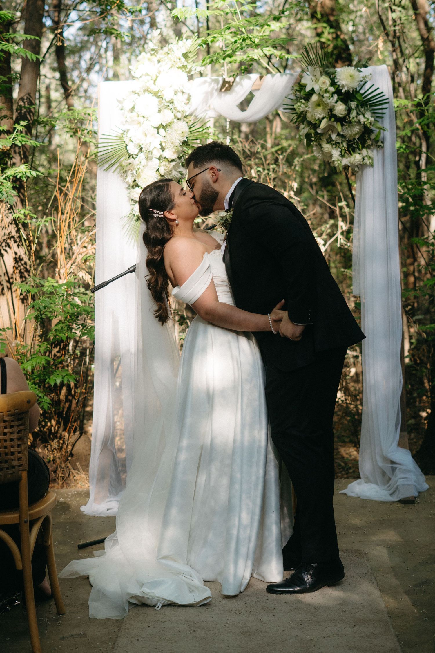 Romantic outdoor wedding ceremony under white draped arch adorned with greenery in woodland setting.