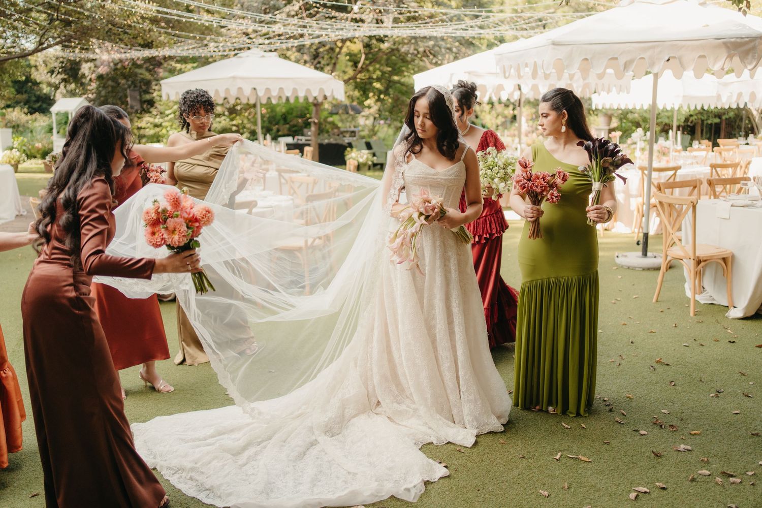 Bridesmaids in green and brown dresses help adjust a bride's long veil at an outdoor garden wedding.