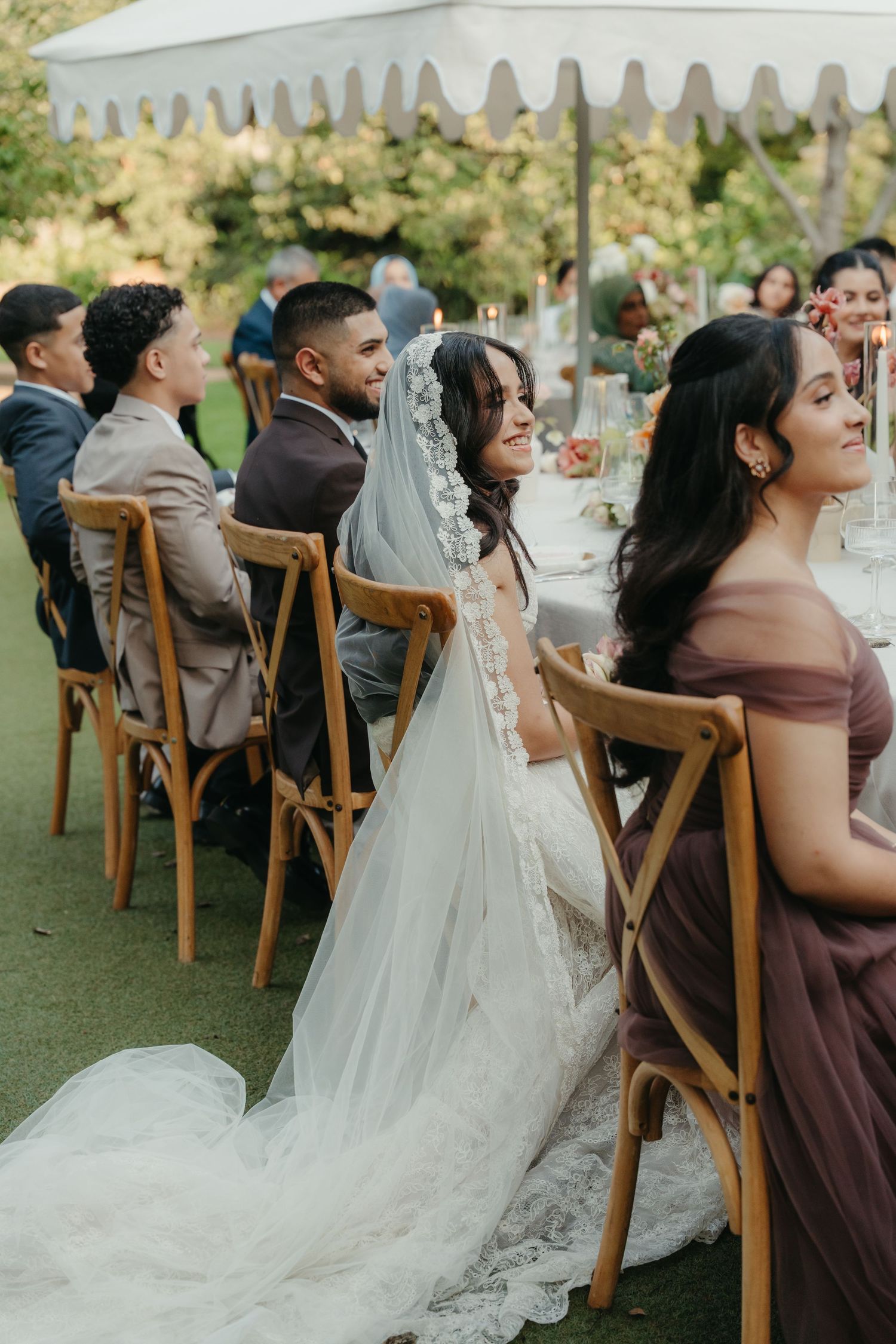 Bride in lace gown and veil sits with groom and guests at outdoor wedding reception table.