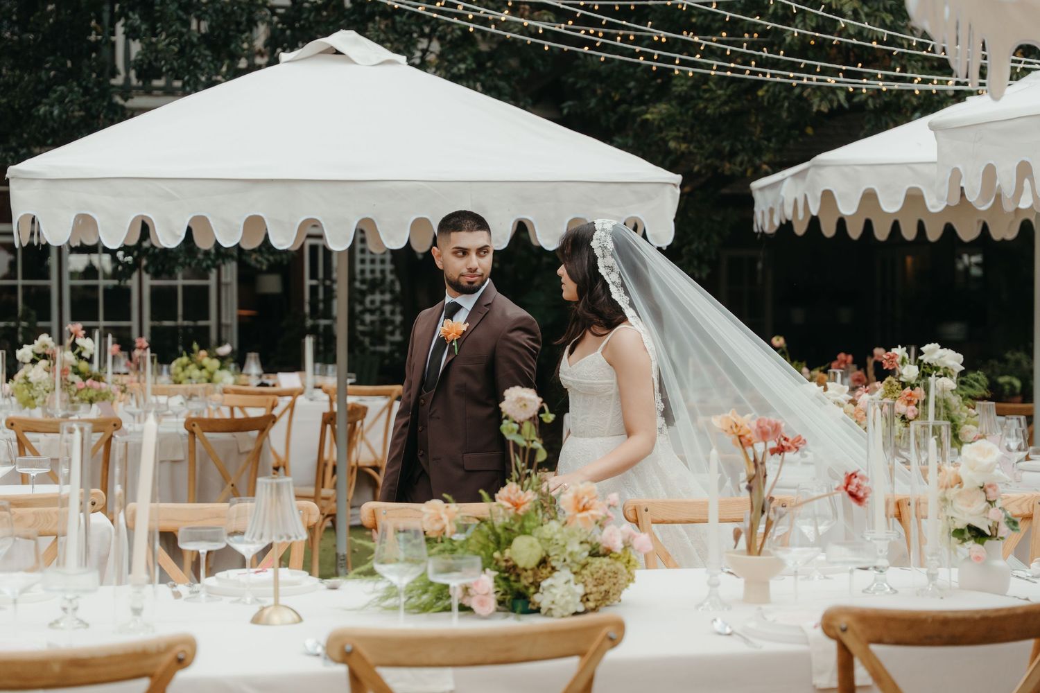 Bride and groom stand among elegantly set outdoor wedding reception tables with floral centerpieces and white tent canopies.