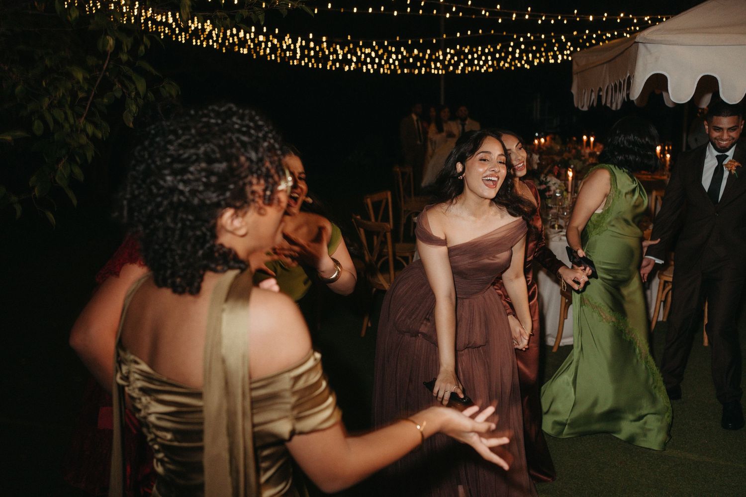 Women in elegant gowns dancing joyfully at an outdoor wedding reception under twinkling string lights at night.
