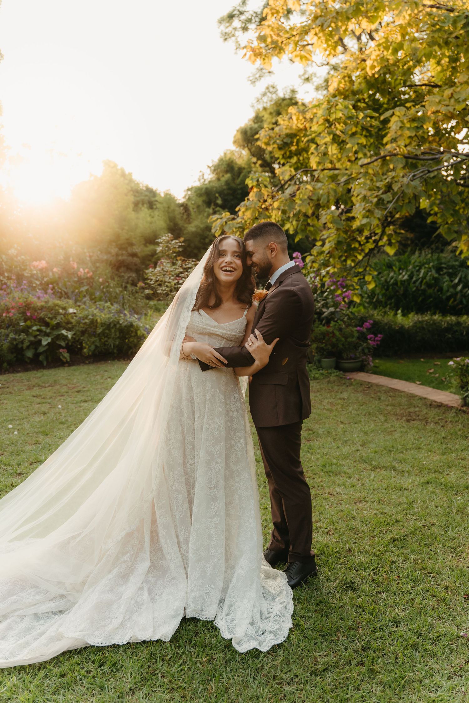 Bride in lace gown and veil embraces groom in brown suit at golden hour in a lush garden wedding.