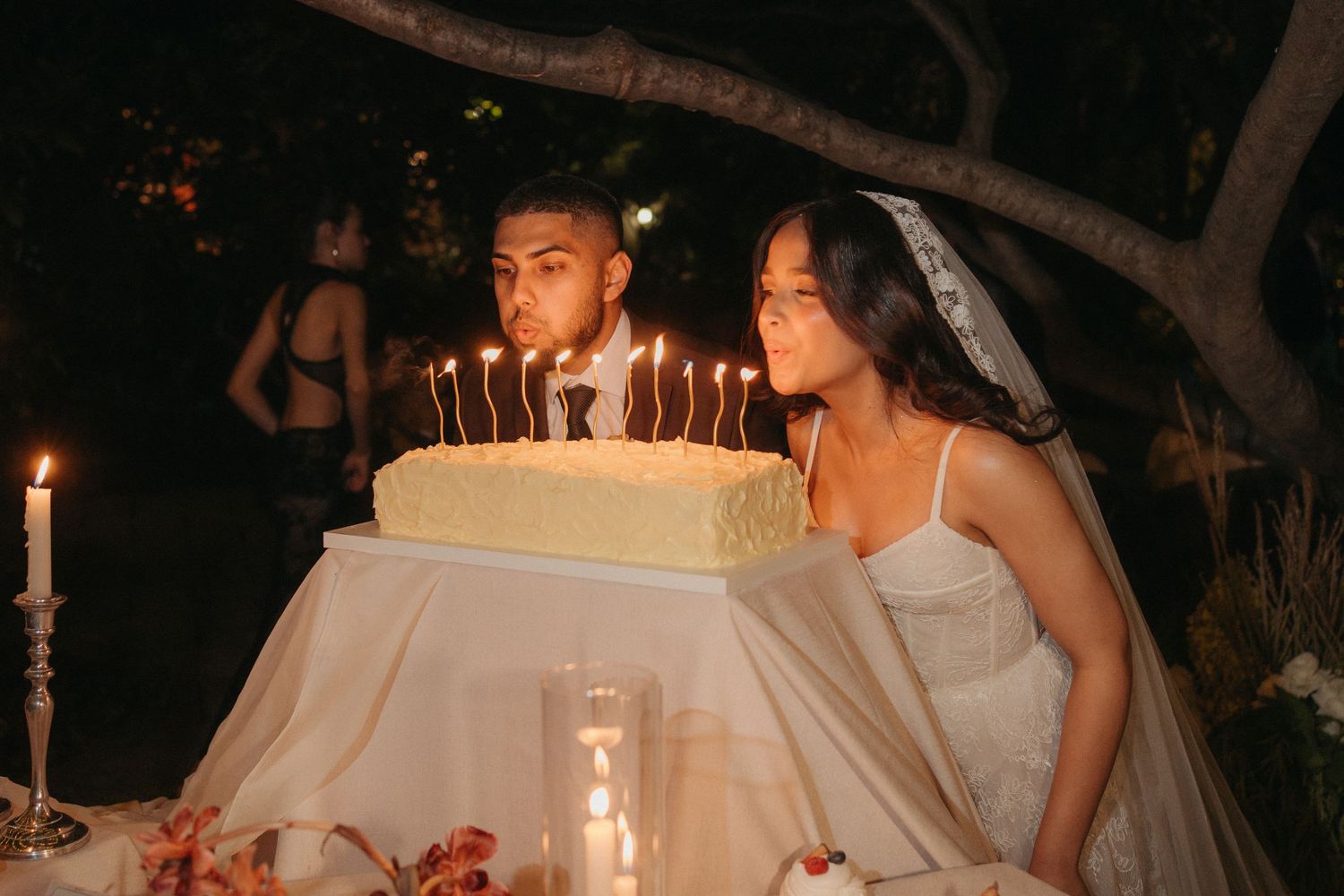 Bride and groom blowing out candles on wedding cake outdoors at night, romantic candlelit ceremony celebration.