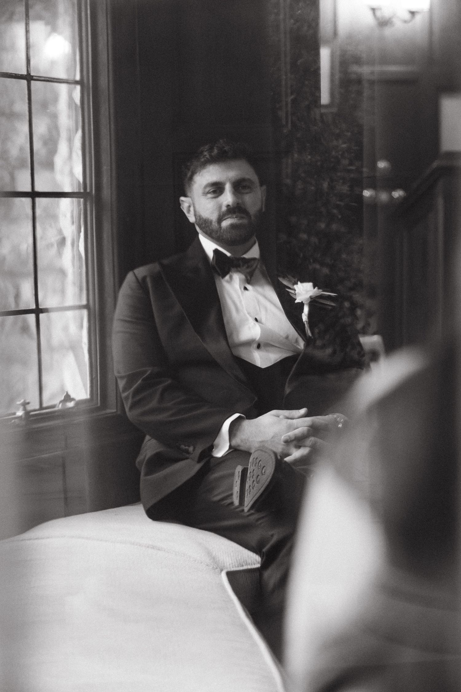 Black and white portrait of a bearded groom in tuxedo with boutonniere seated by a window.