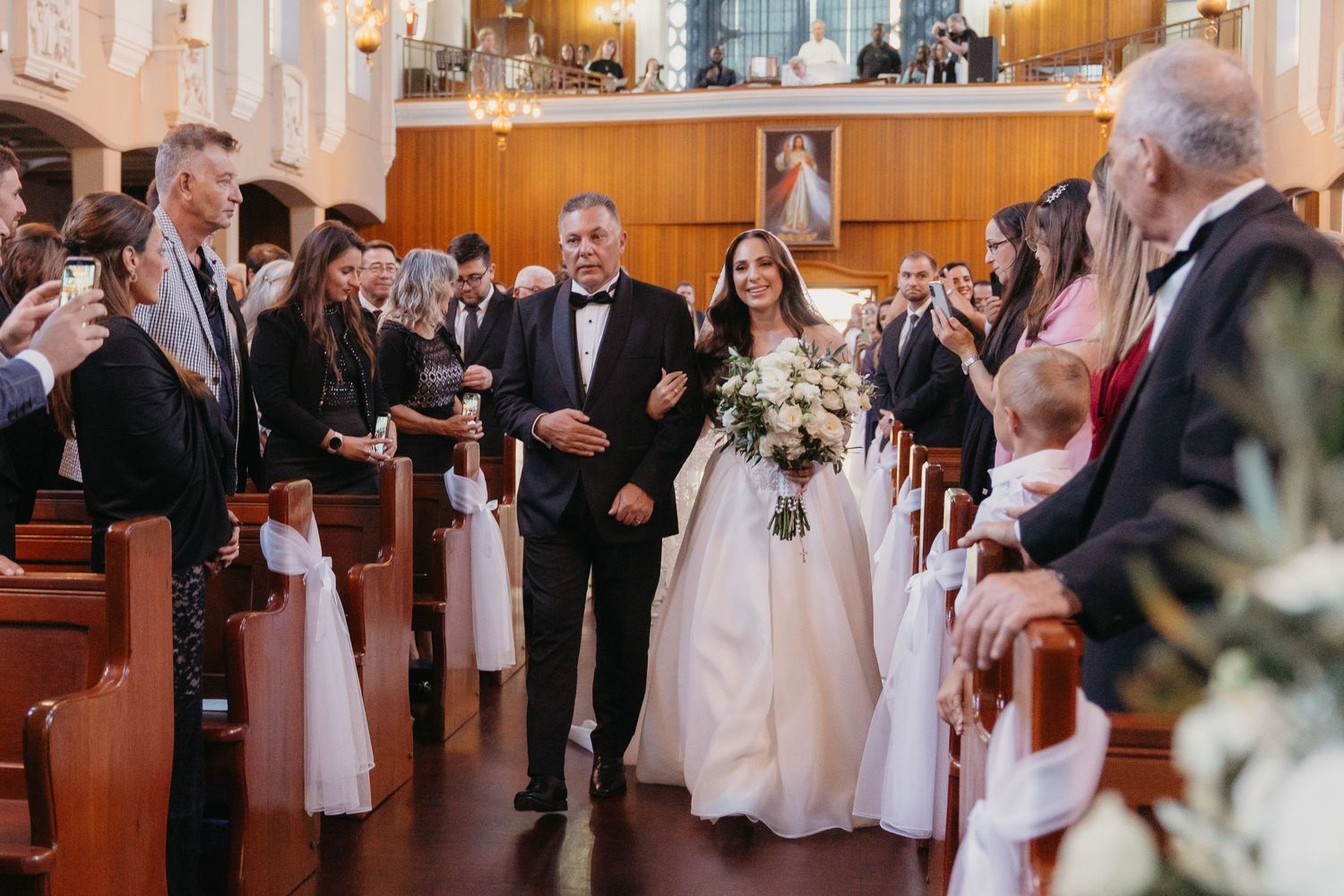 Bride walks down Rosebank Catholic Church aisle with father during wedding ceremony, guests standing in pews, holding bouquet.