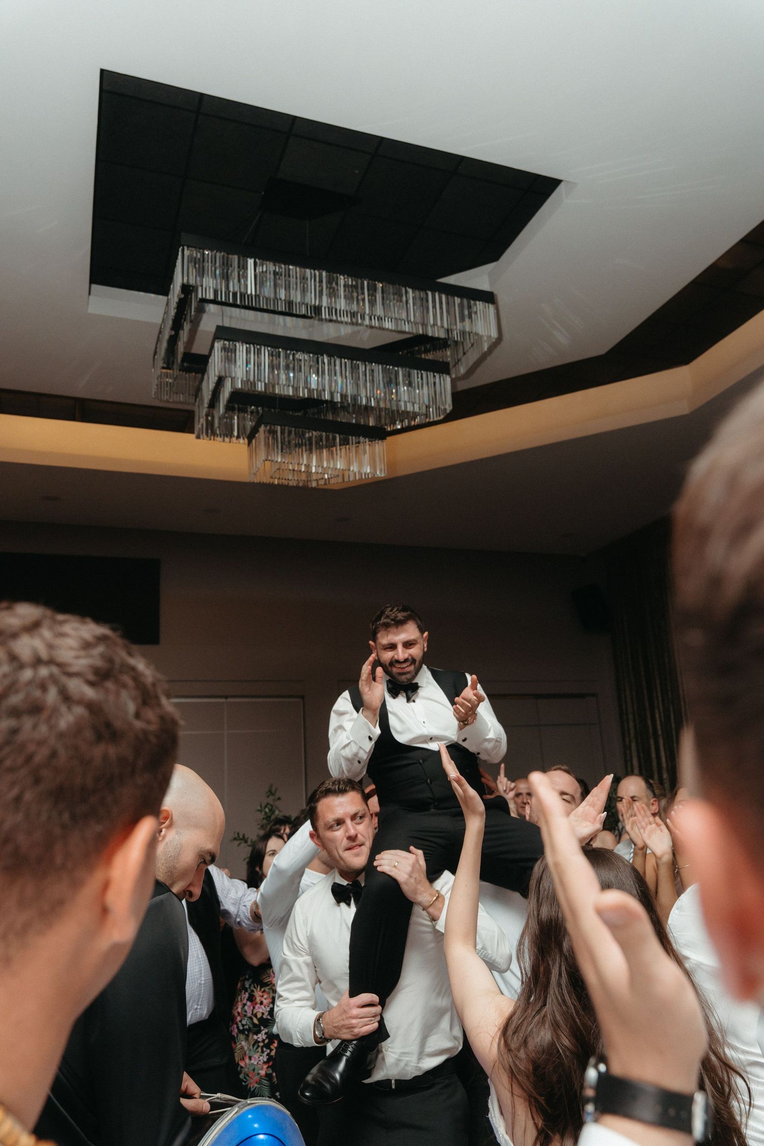 Groom lifted on shoulders by groomsmen during lively wedding reception celebration in elegant ballroom.