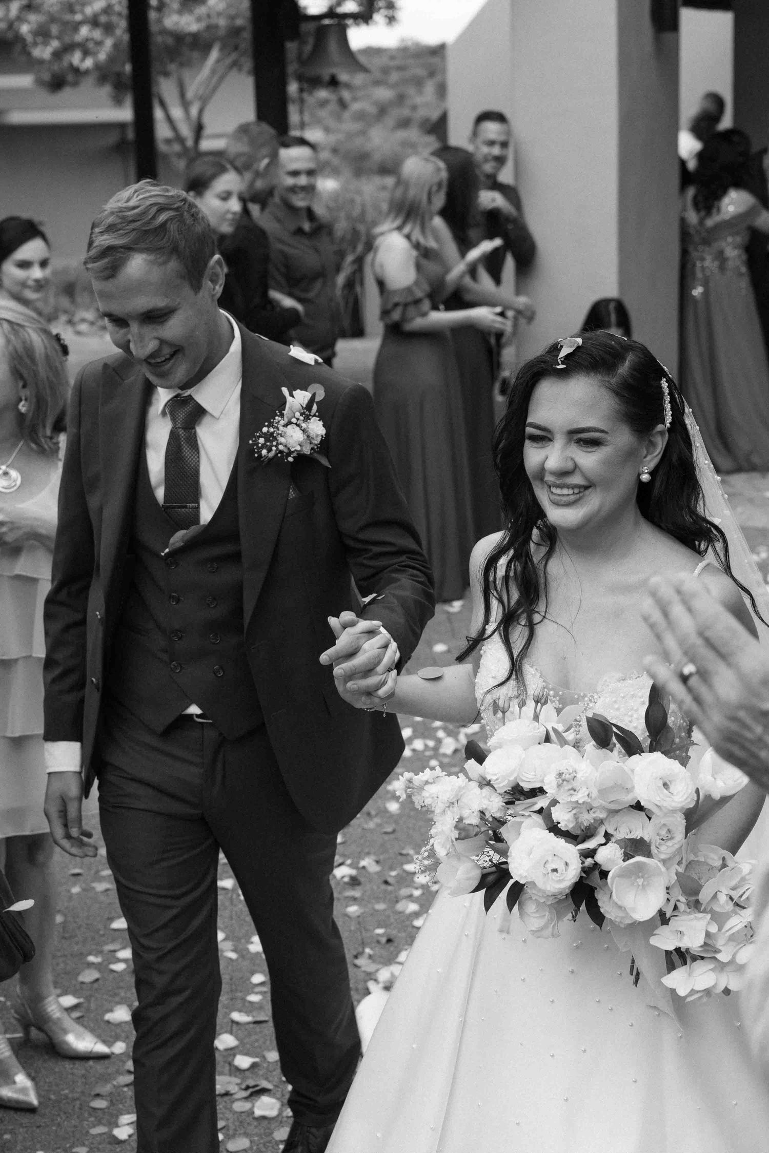Black and white photo of a smiling bride holding flowers and groom in suit walking together at their wedding ceremony.