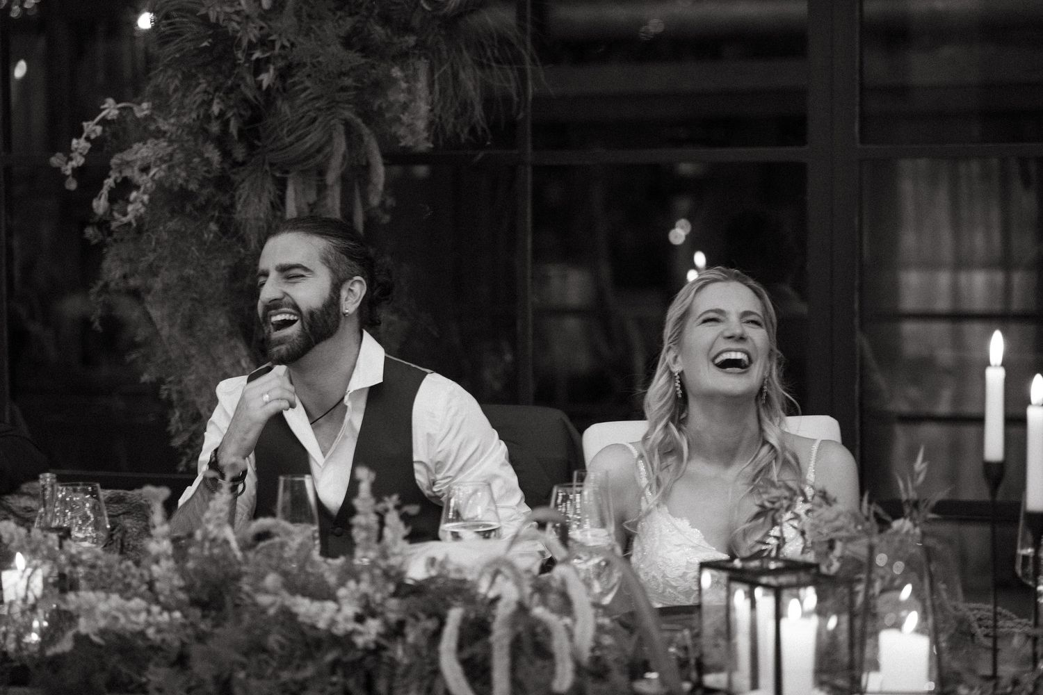 Black and white photo of a laughing bride and groom seated at their wedding reception table at Rosemary Hill with candles and florals.