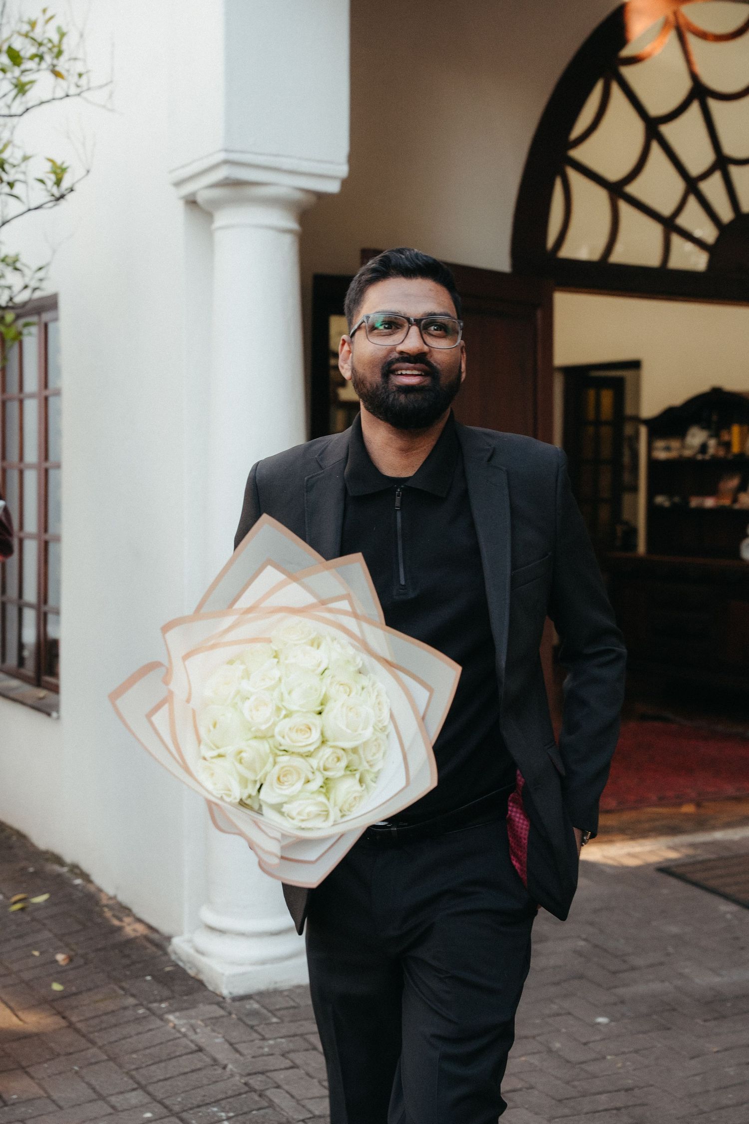 Smiling bearded man in black suit holding large white rose bouquet outside elegant white building with arched entrance.