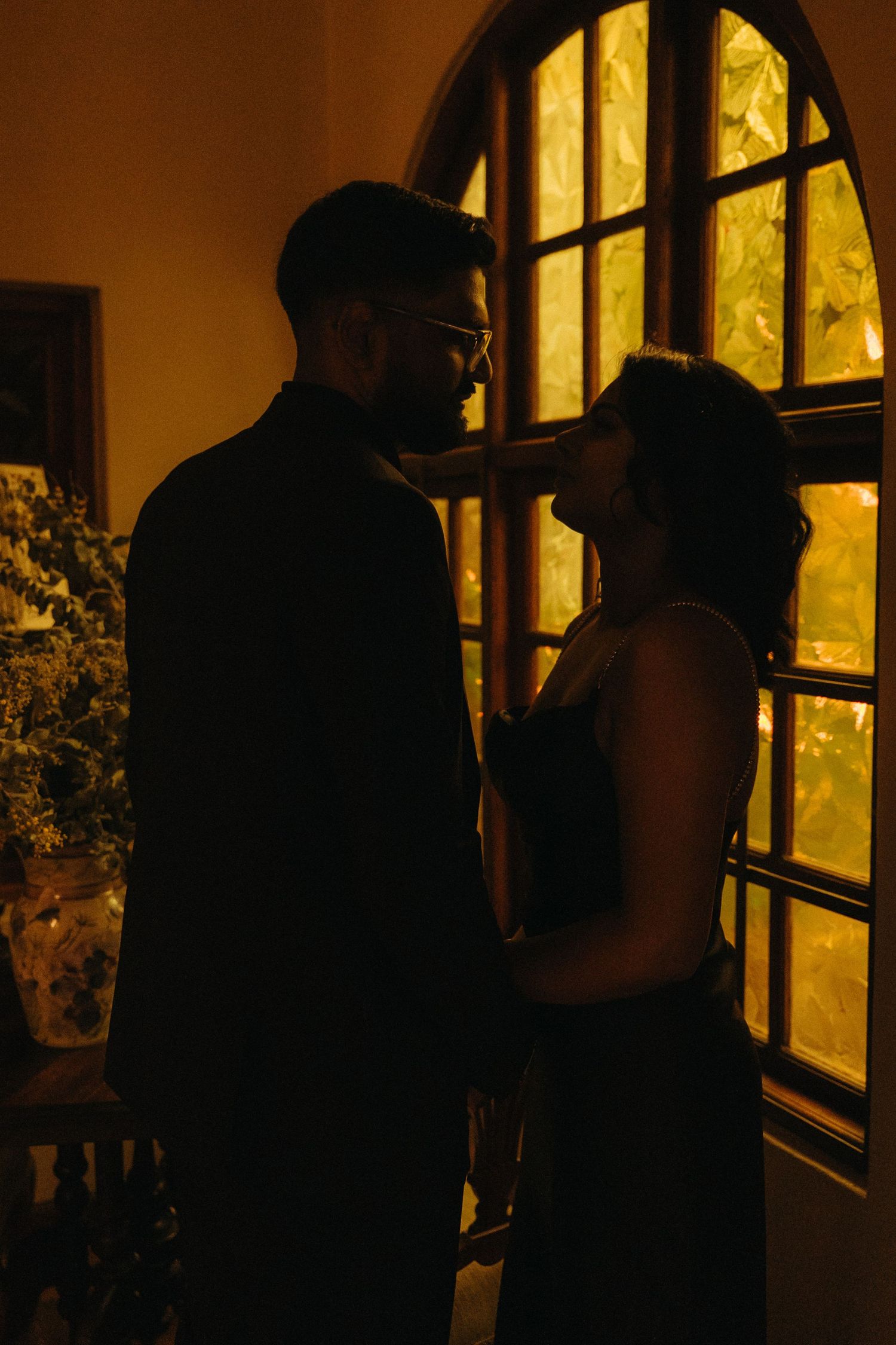 Romantic couple silhouetted against warm glowing arched window in dimly lit room.