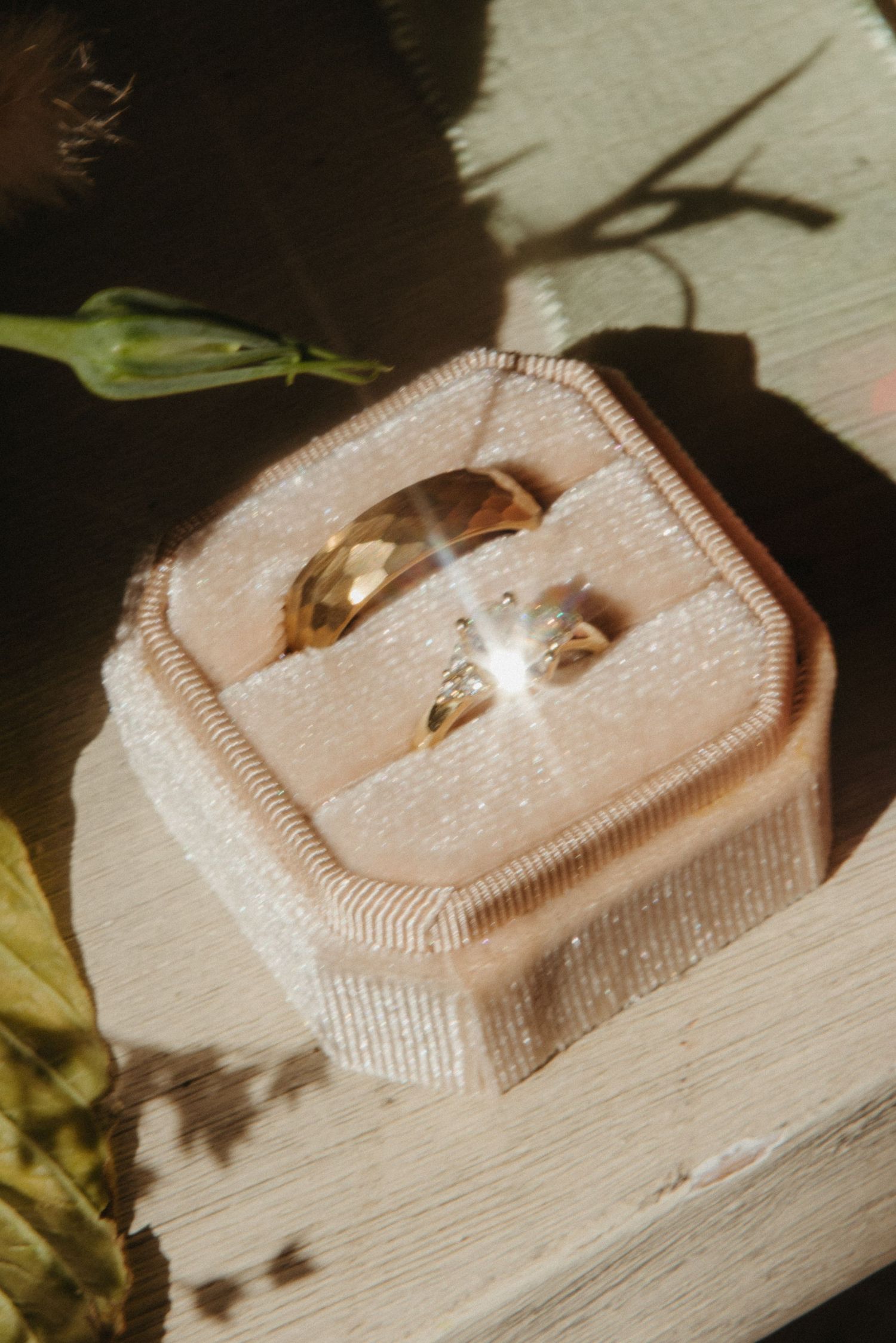 Two gold wedding rings in a pink velvet octagonal ring box, sunlit with floral shadows.