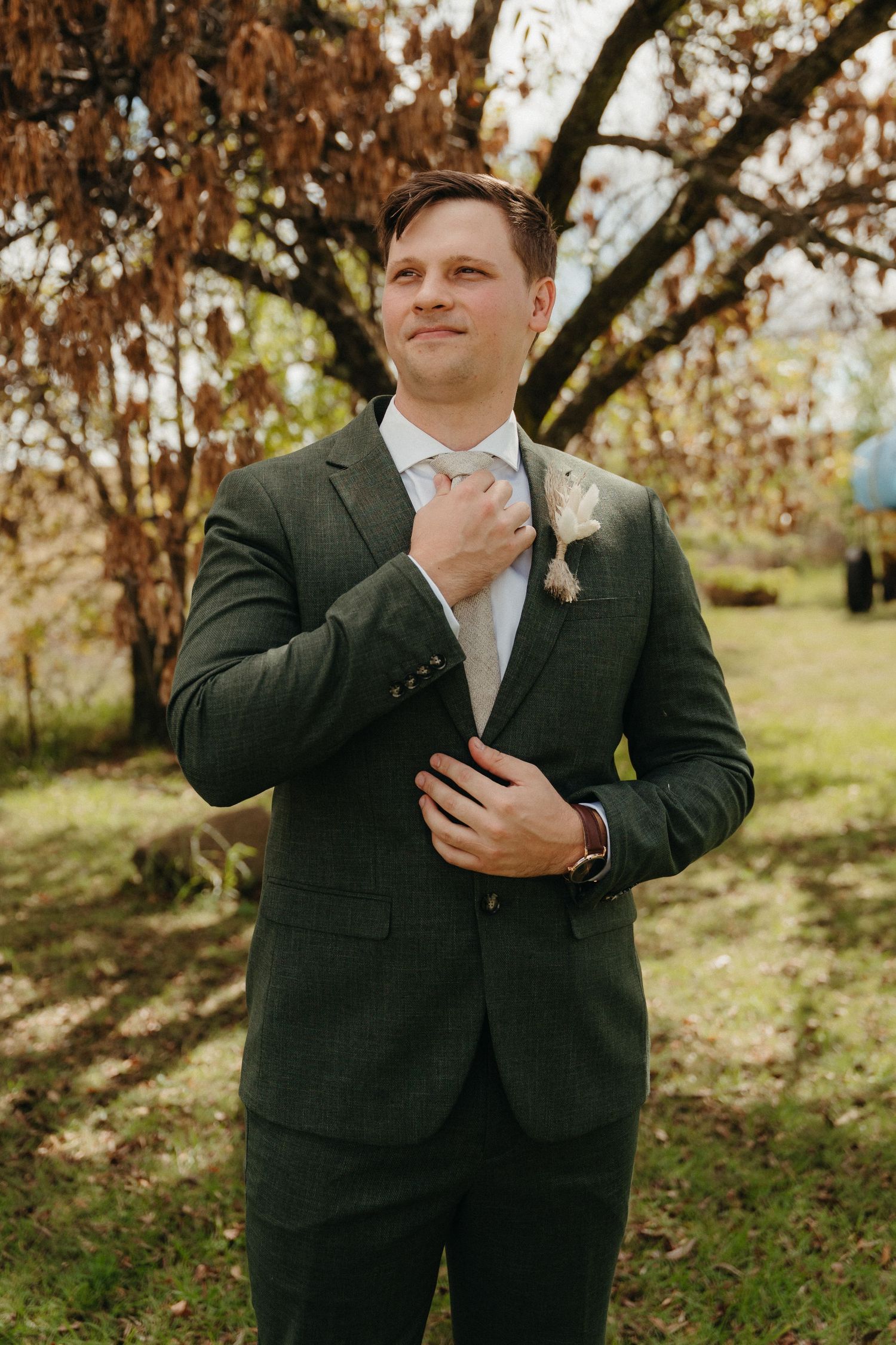 Groom in dark green suit adjusting tie outdoors with autumn trees in background.