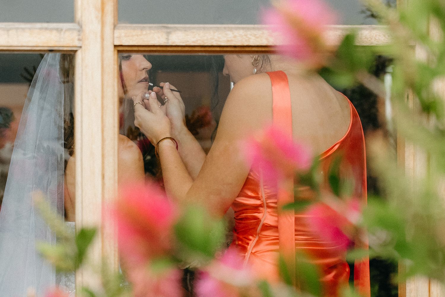 Bridesmaid in orange satin dress helping bride with veil near window, pink flowers in foreground.