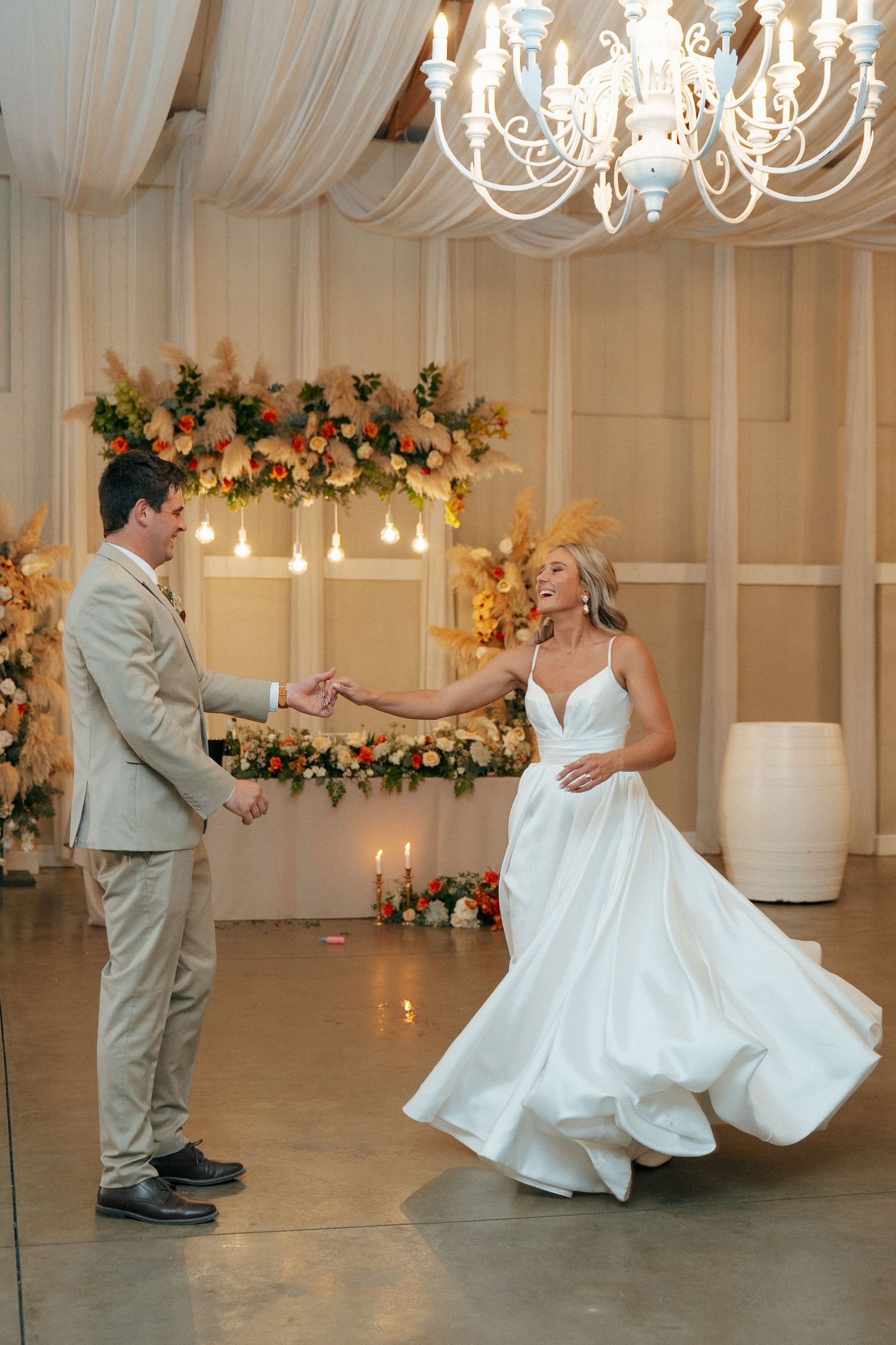 Bride in a flowing white gown twirls during her first dance with the groom in a beige suit beneath a chandelier and floral arch.