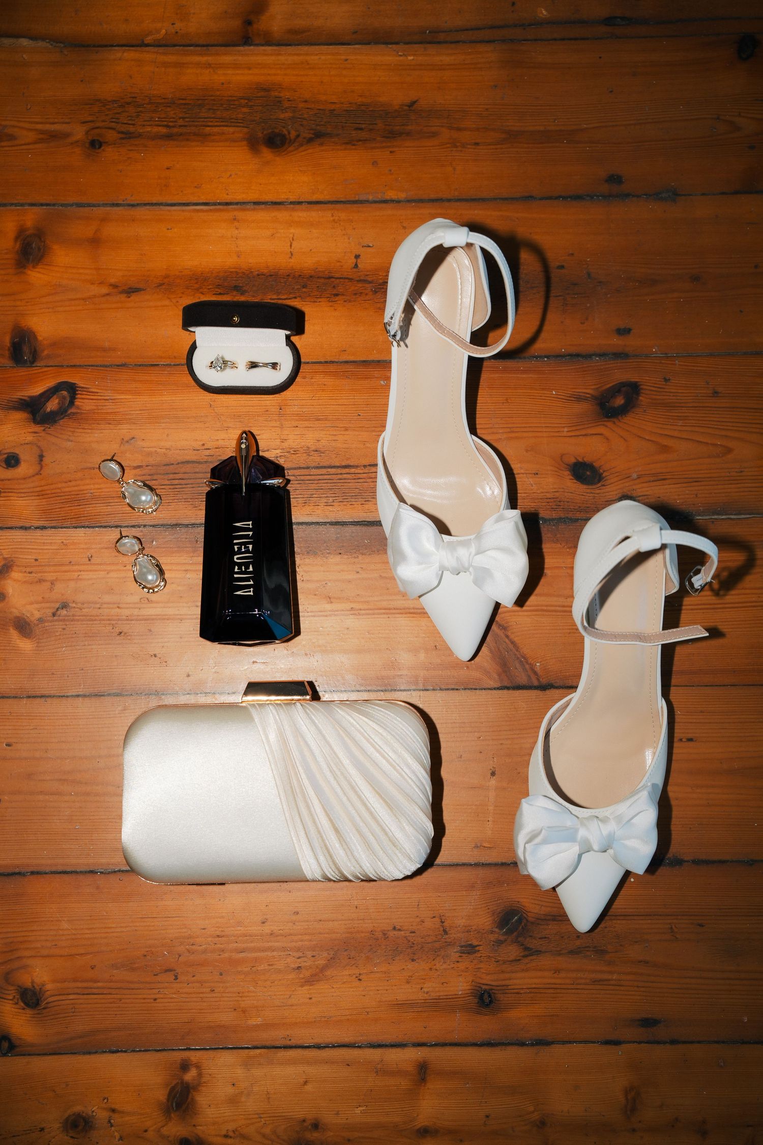 Flat lay of bridal accessories on a wooden floor with white bow heels, a satin clutch, perfume bottle, drop earrings and a ring box.