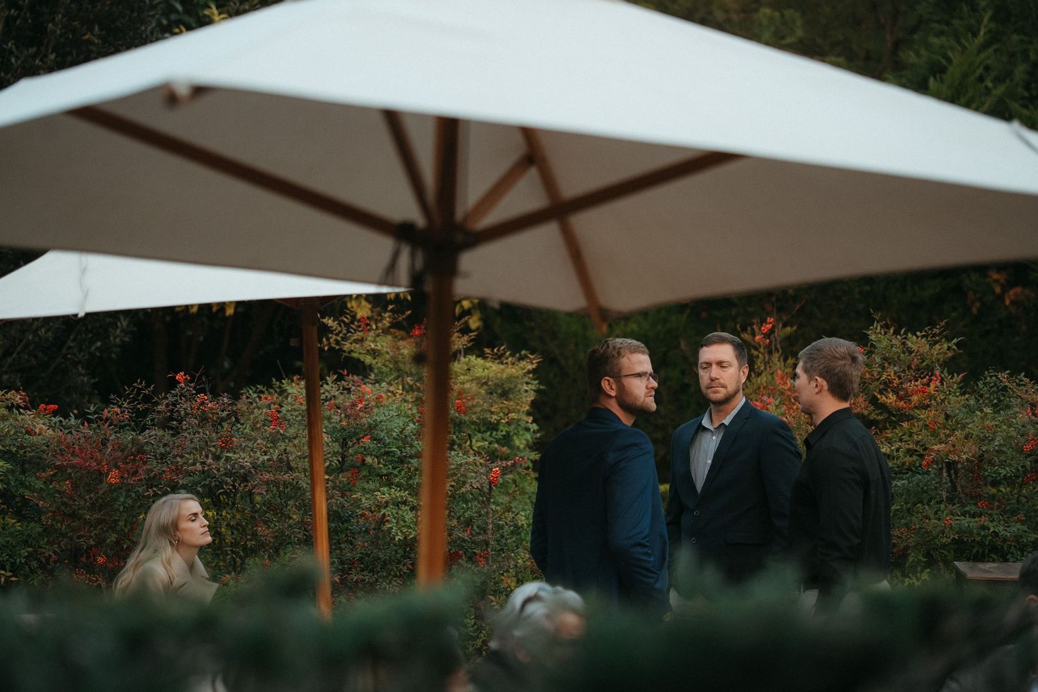 Four people conversing outdoors under a white patio umbrella surrounded by lush greenery at an evening garden event.