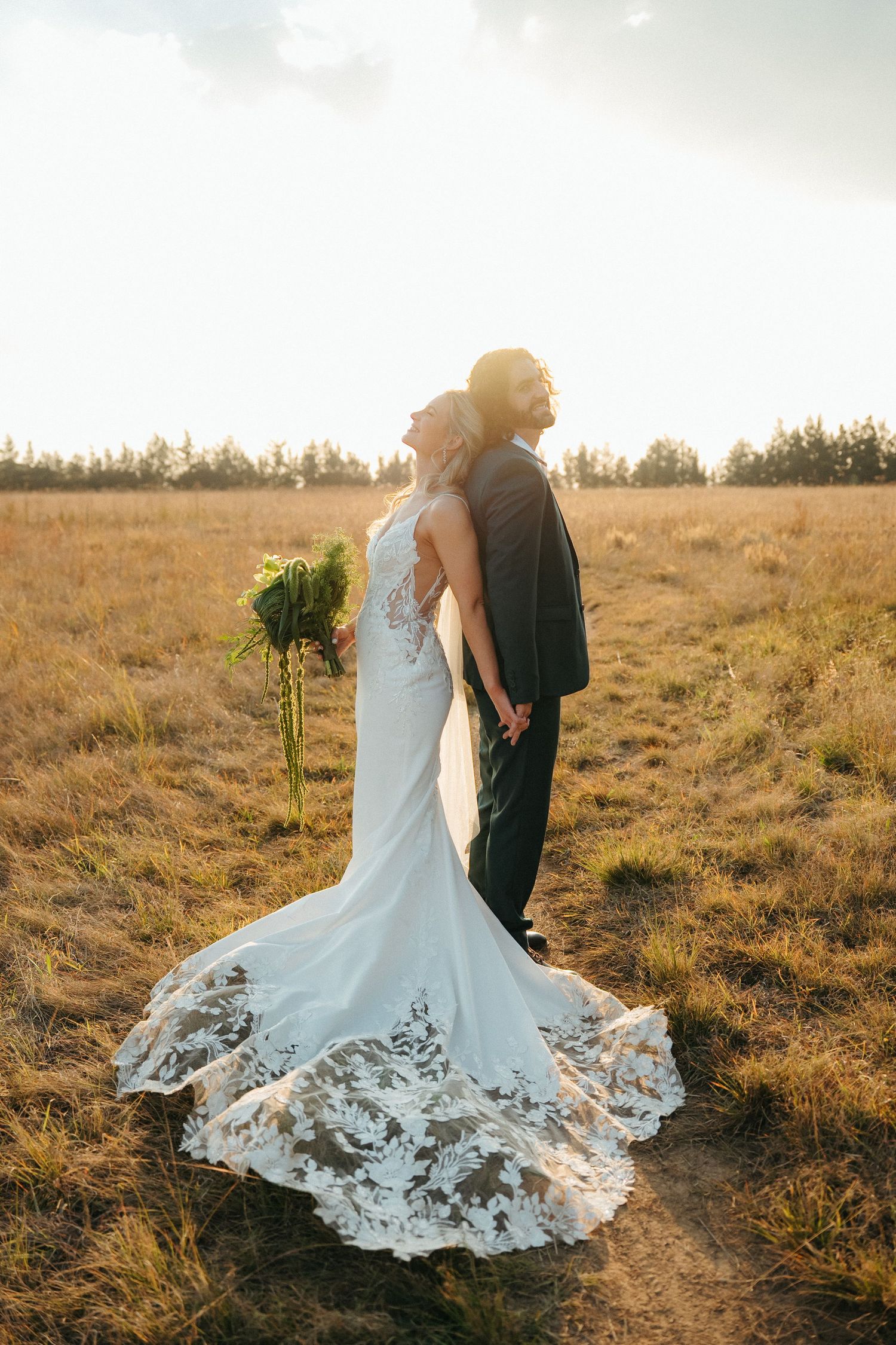 Bride and groom standing back-to-back in golden field at sunset, bride in lace train gown holding bouquet.