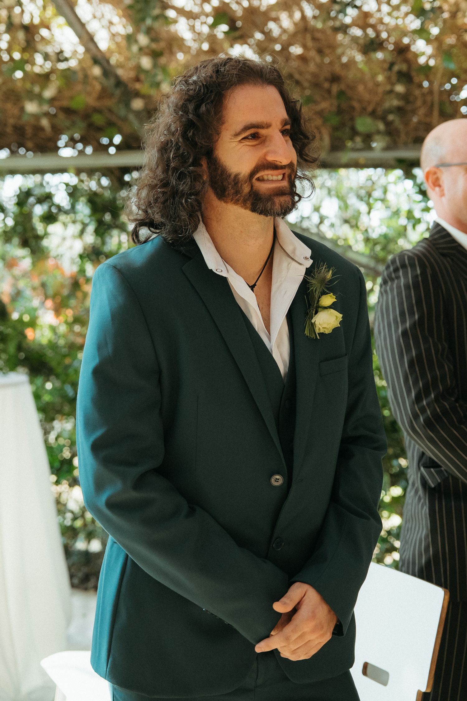 Smiling groom in dark green suit with floral boutonniere stands at wedding ceremony altar with lush greenery backdrop.