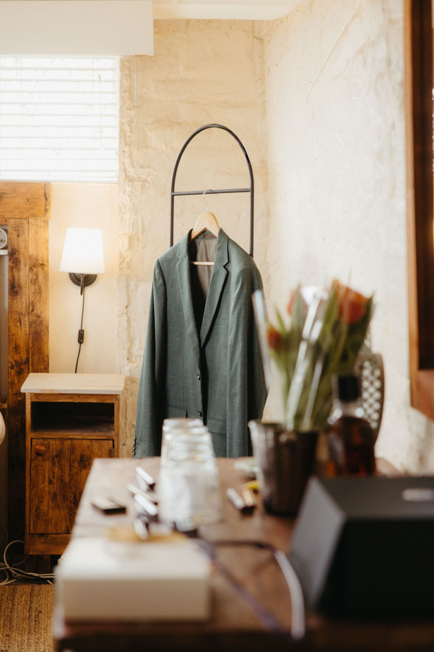 Green blazer on a rack in a rustic room with wooden furniture, a lamp, and floral arrangement on a desk.