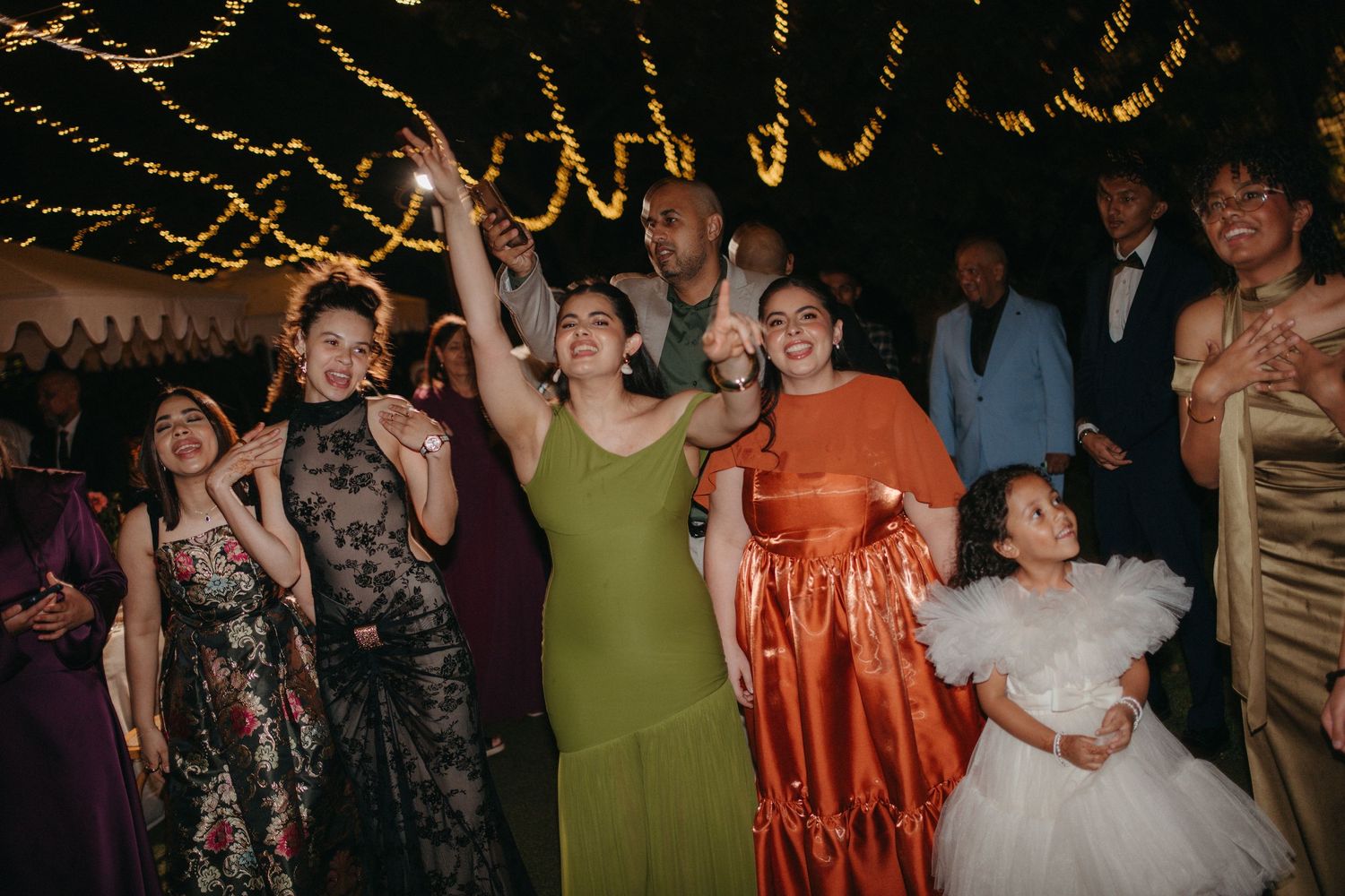 Joyful wedding guests dancing and celebrating at a festive evening reception with warm string lights overhead.