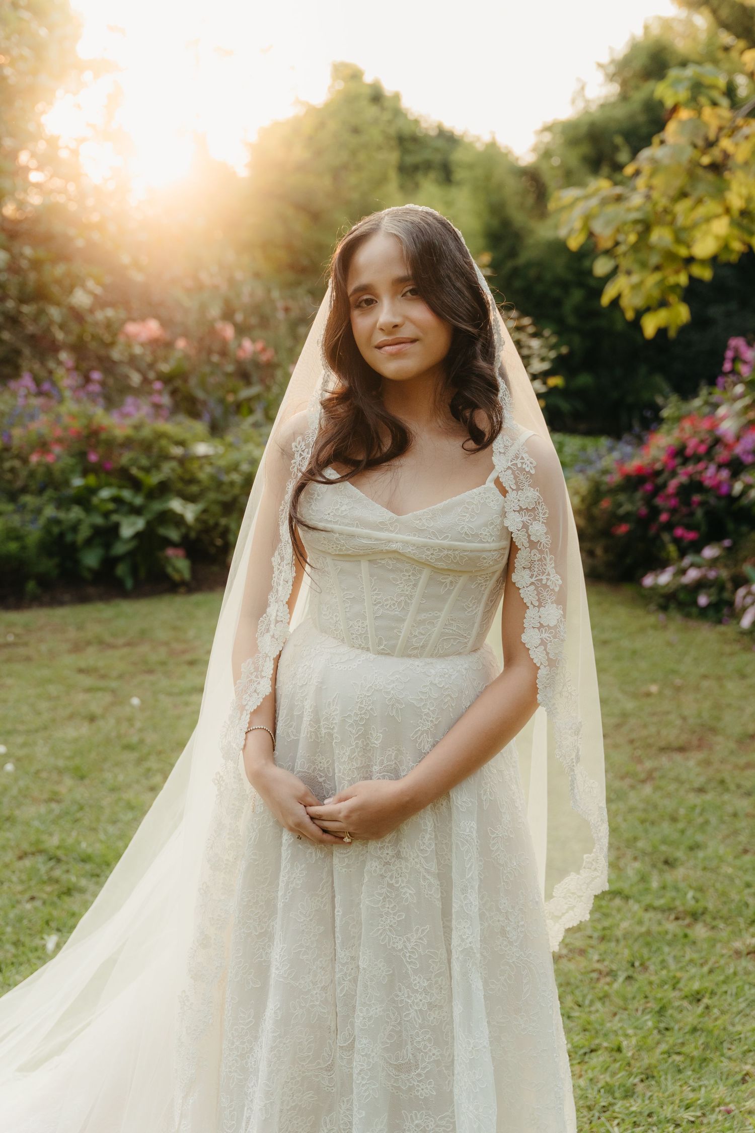 Bride in lace wedding dress and veil poses in sunlit garden with colorful flowers at golden hour.