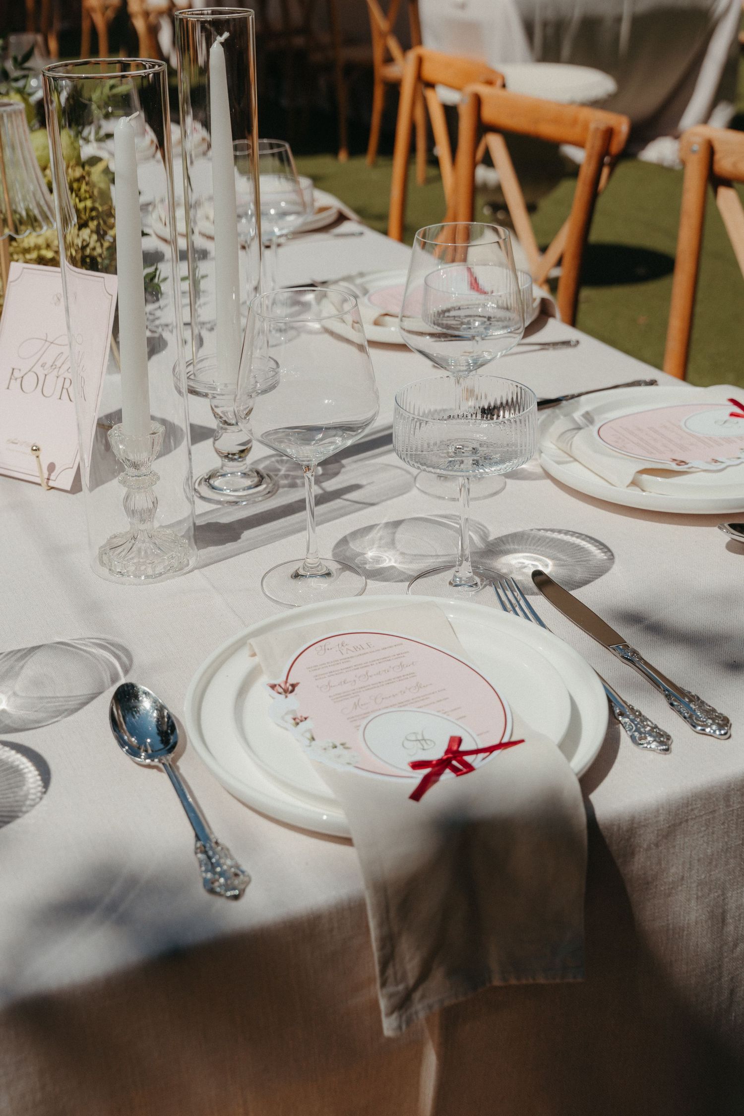 Elegant wedding table setting with crystal glasses, white plates, red bow menus, candles, and linen napkins on a white tablecloth.