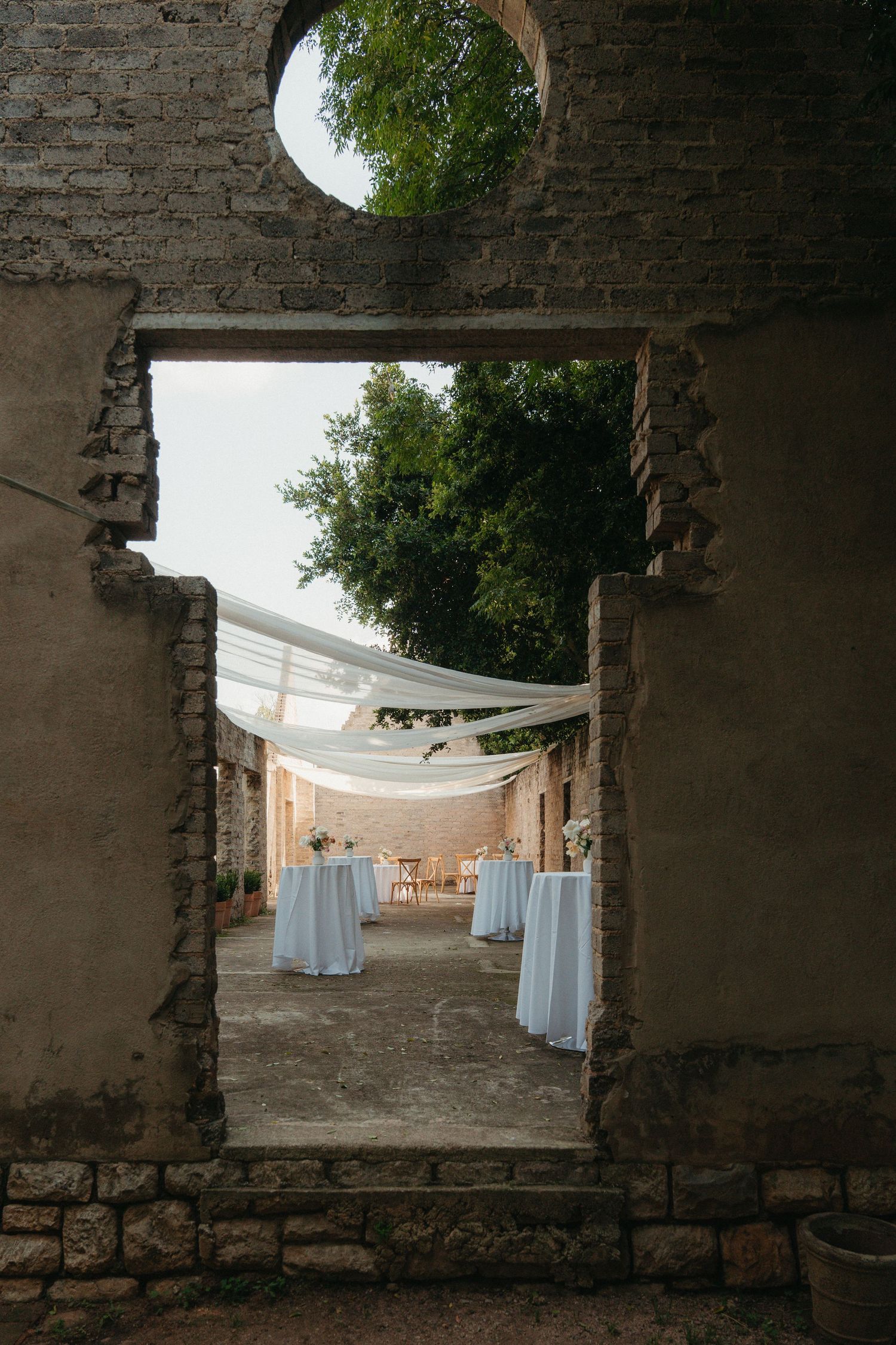 Elegant outdoor wedding reception setup viewed through rustic stone archway with white draped tables.