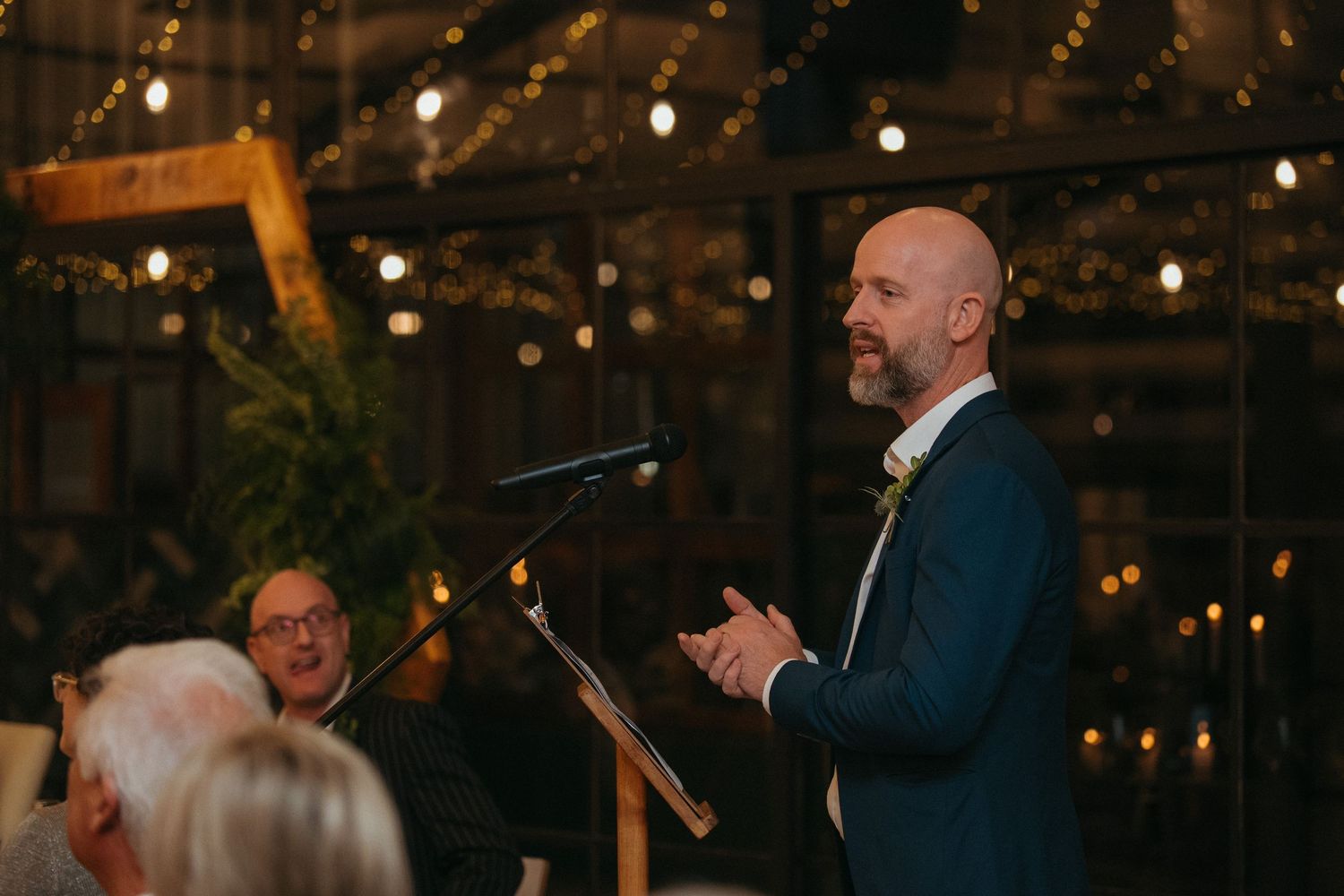 Bald man in blue suit giving a speech at an elegant wedding reception with warm string lights.