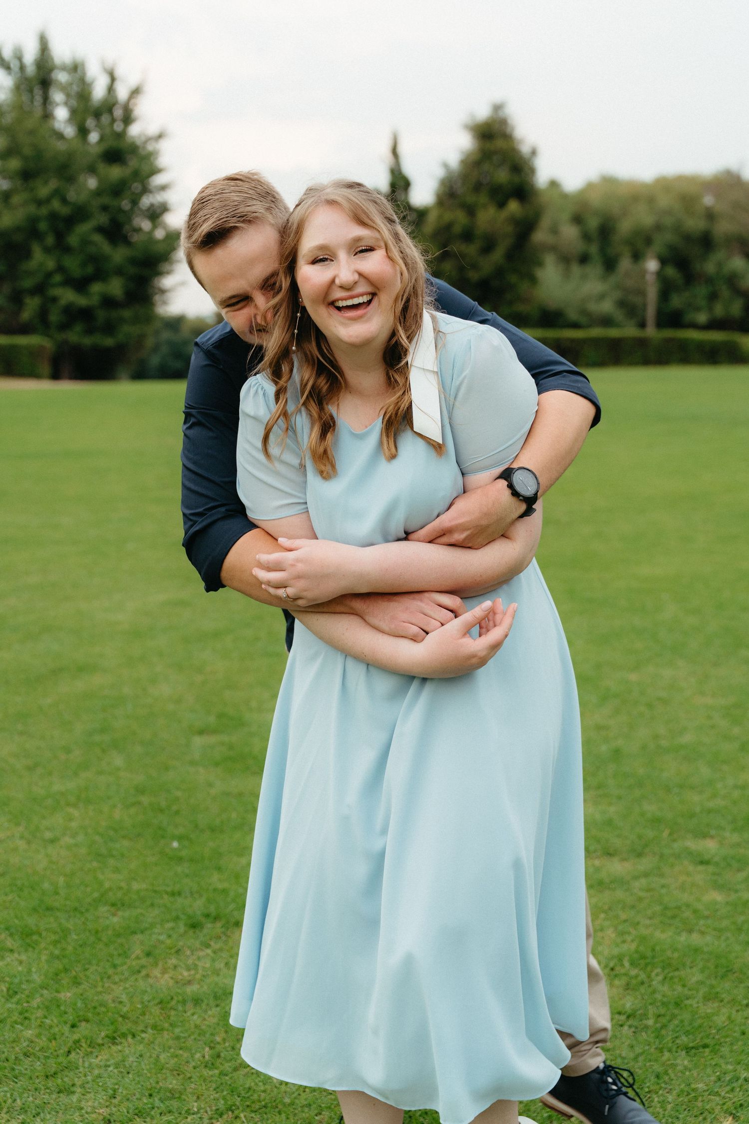 A couple embraces playfully in a green field on a sunny day, the person in a light blue dress laughing joyfully.
