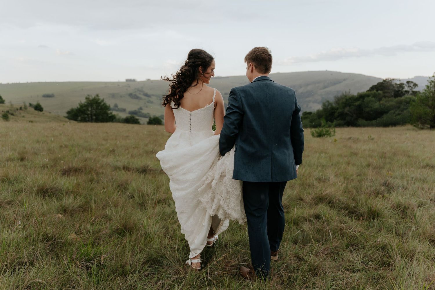 A couple walks hand in hand through a scenic hillside meadow on their wedding day.