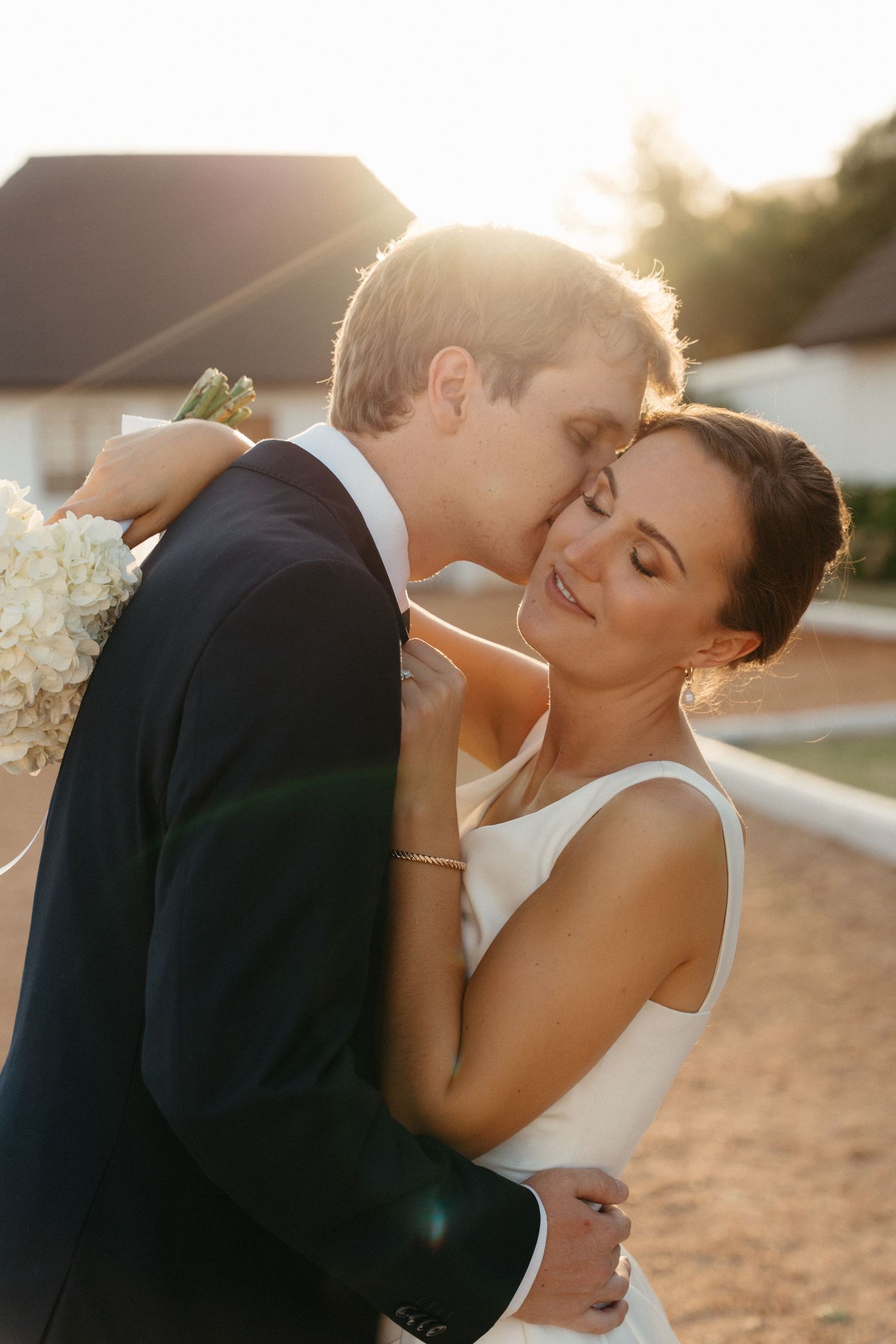 A romantic sunset wedding photo of a couple embracing outdoors with golden light glowing behind them.