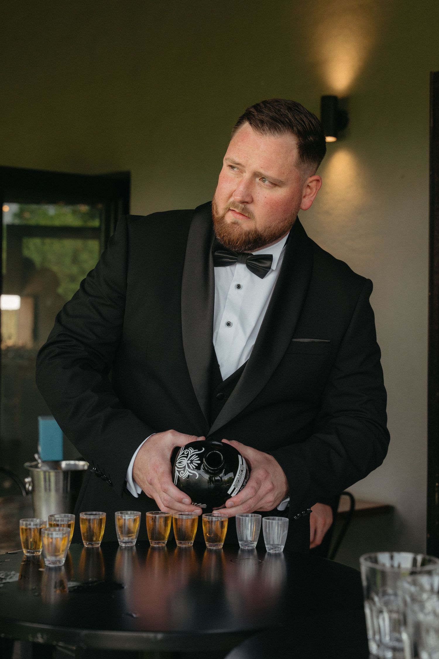 Bartender in black tuxedo carefully arranges and pours a flight of craft beer samples at wooden bar counter.
