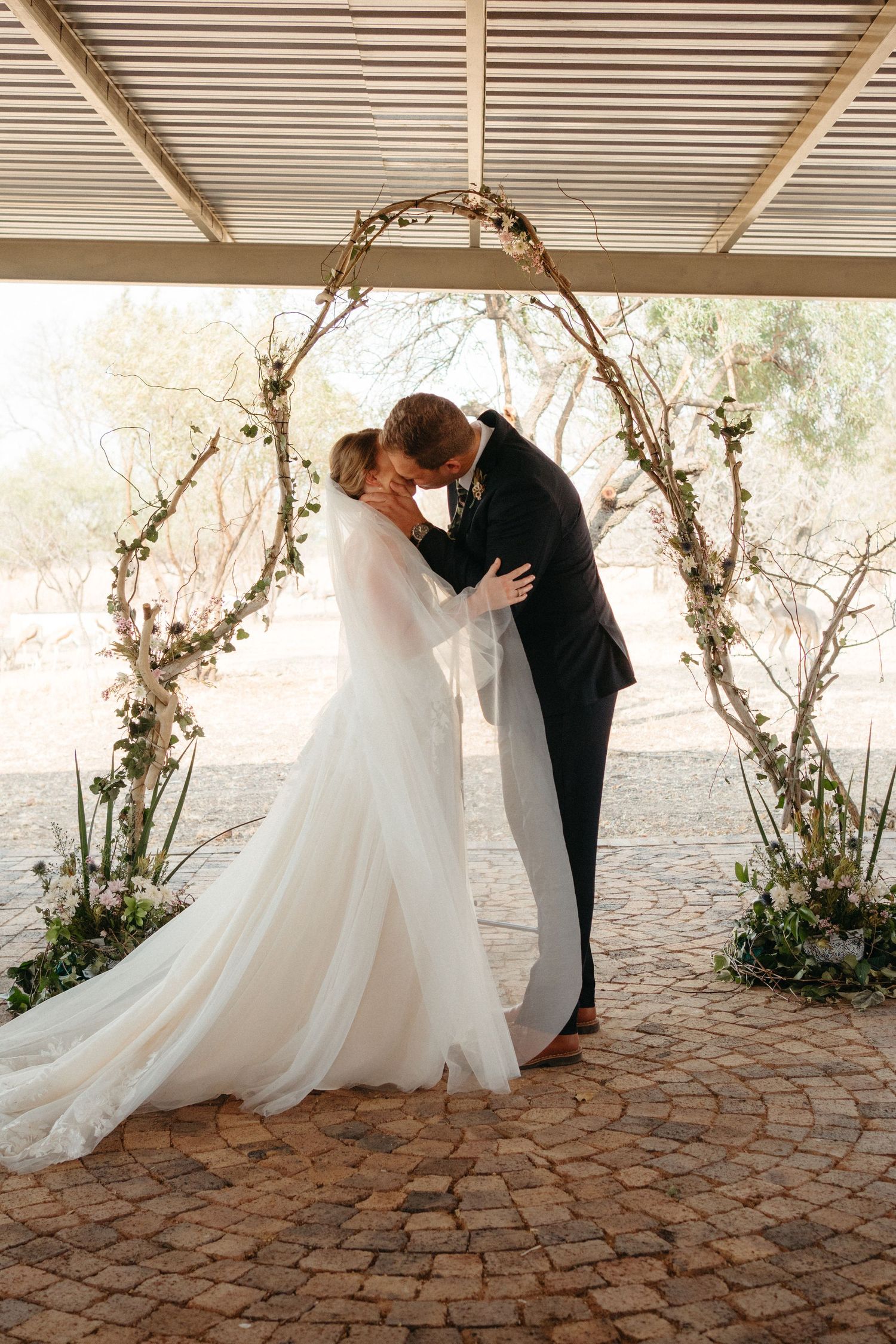 Newlyweds share their first kiss under a romantic floral arch during an outdoor ceremony.