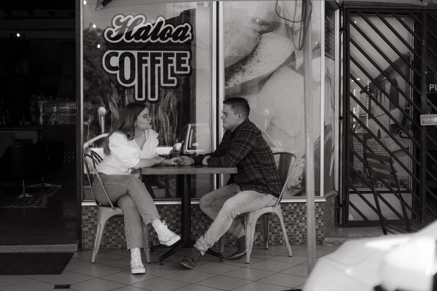 Two people sit at an outdoor table in front of a coffee shop window in a black and white photograph.