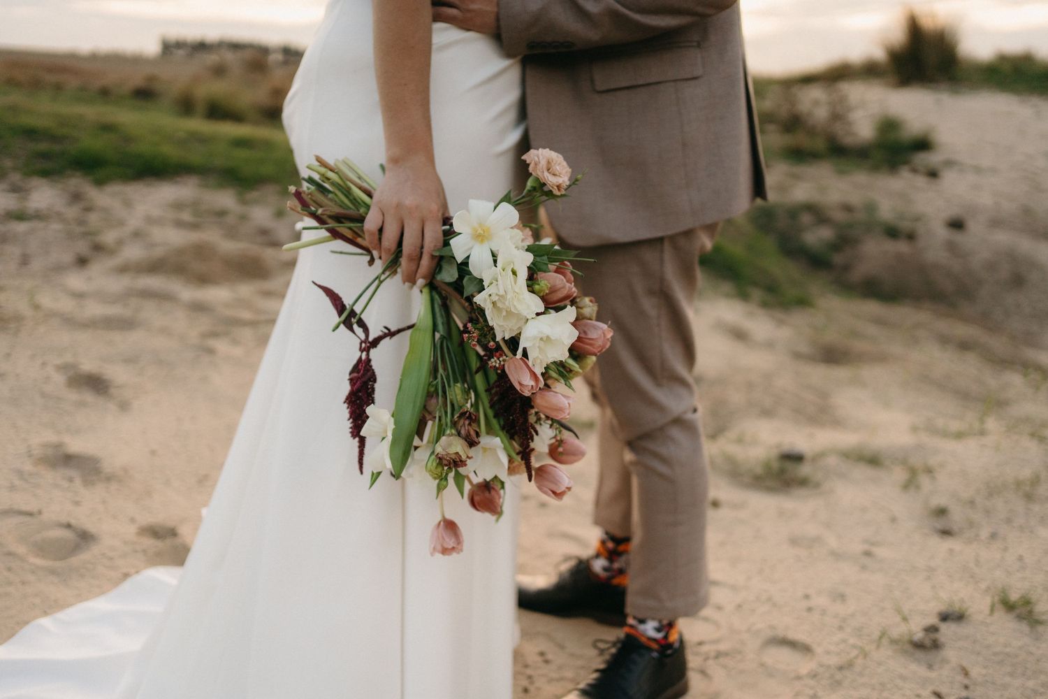 Wedding bouquet of white flowers and greenery held against a white dress and beige suit on a rustic pathway.