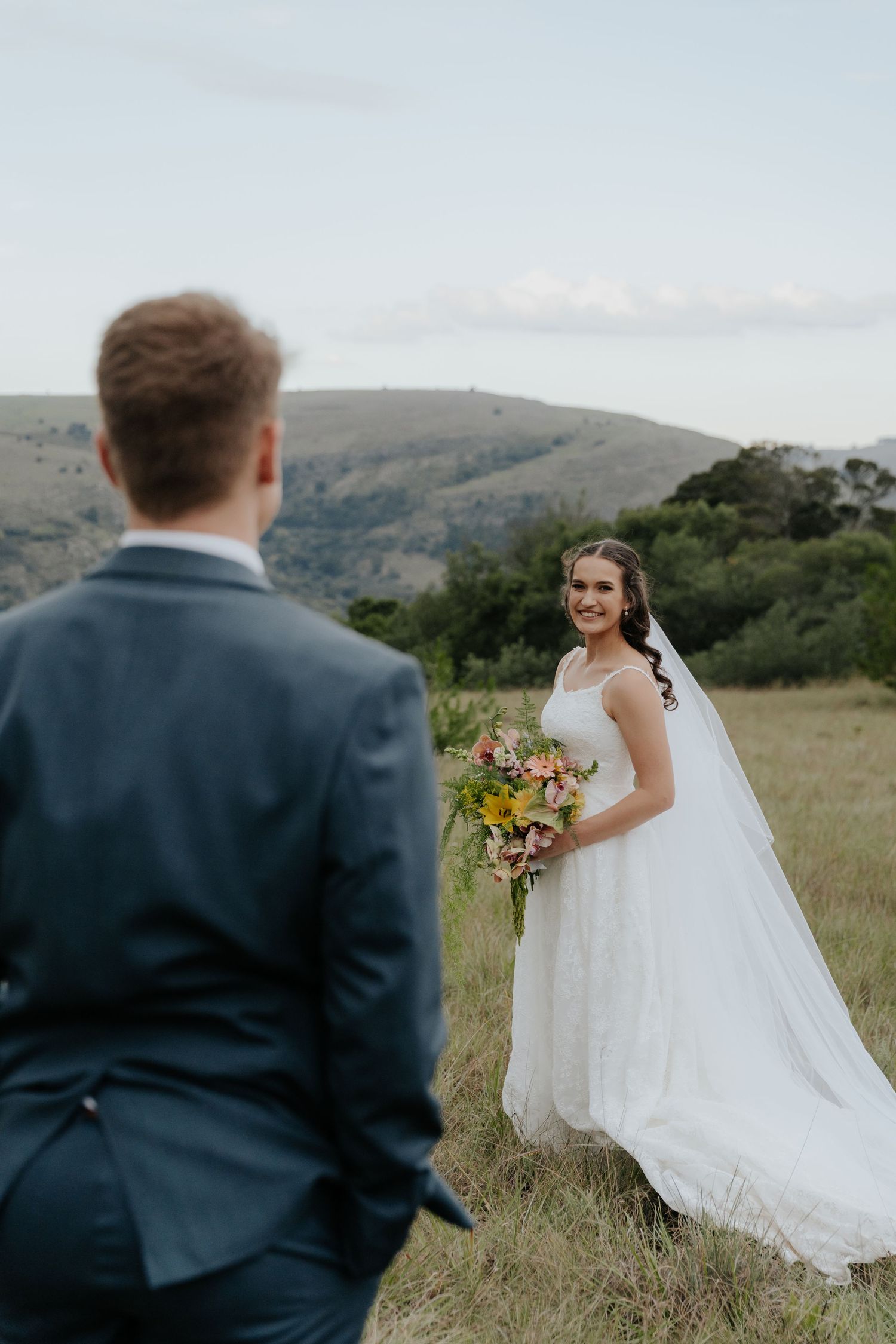 Smiling bride holds colorful bouquet in a scenic outdoor field with rolling hills in Mpumalanga, South Africa