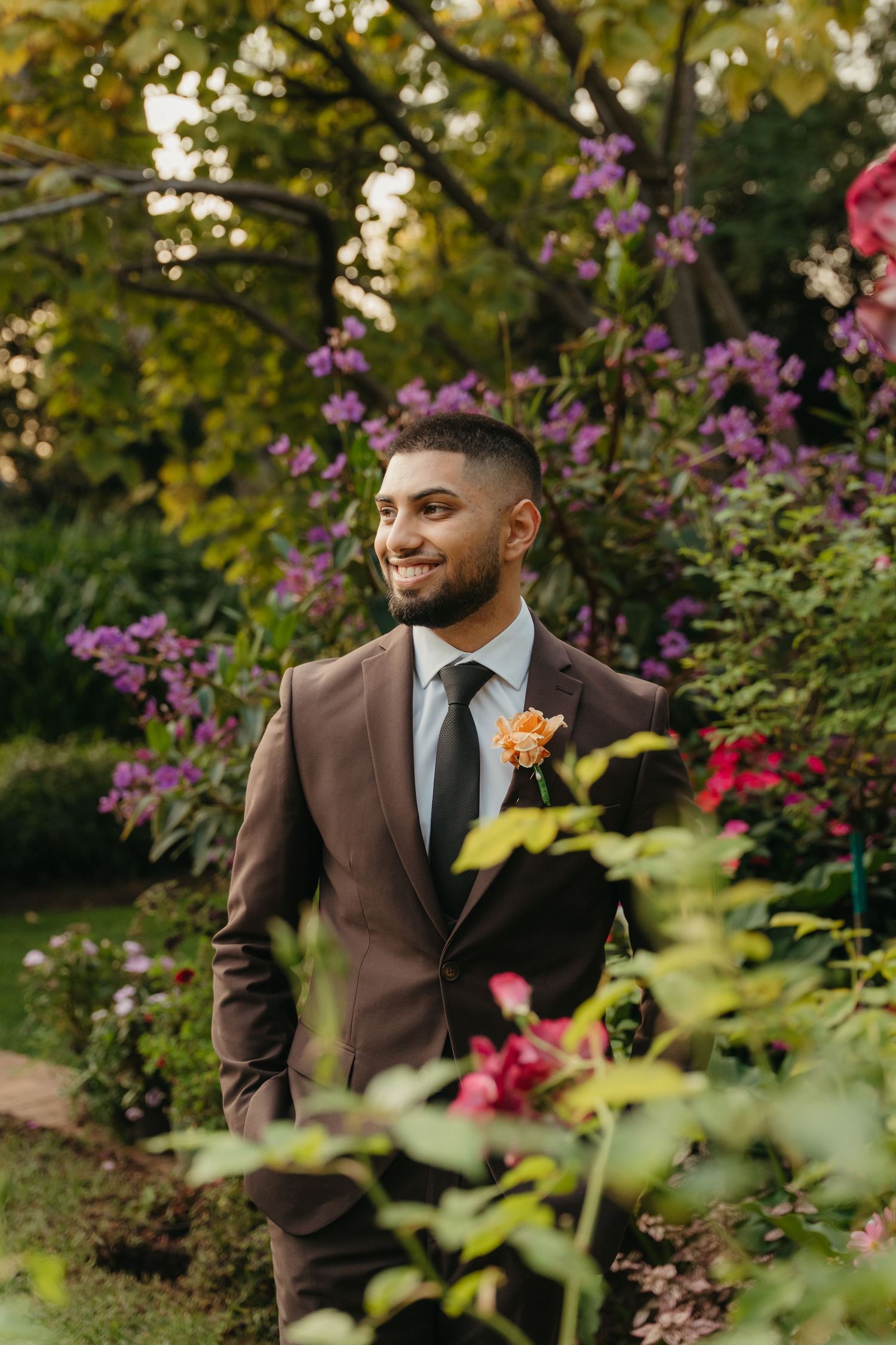 Smiling groom in brown suit with orange boutonniere stands among purple and pink garden flowers at Beechwood Gardens