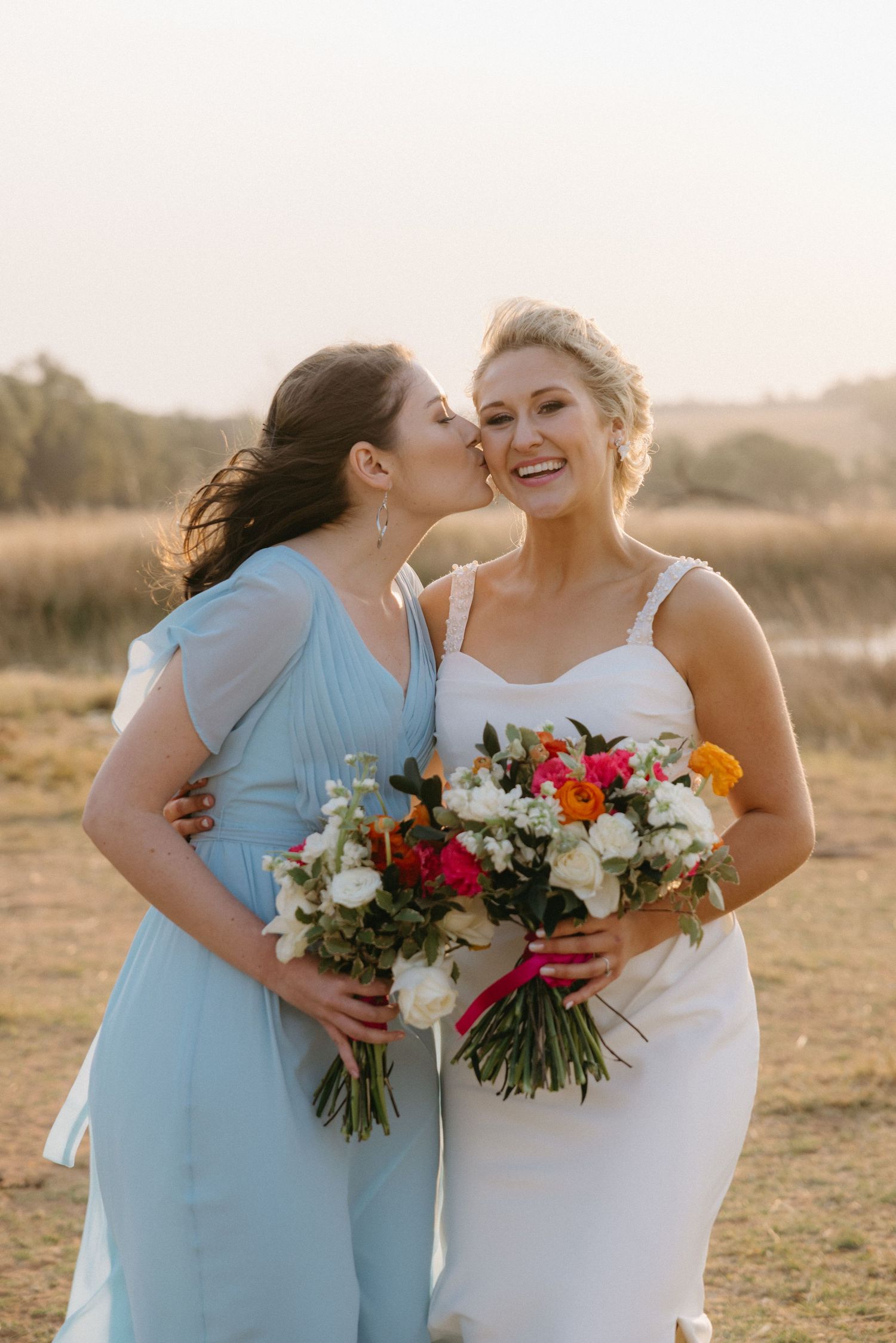 Joyful wedding party members embracing while holding colorful bouquets in a field at sunset.