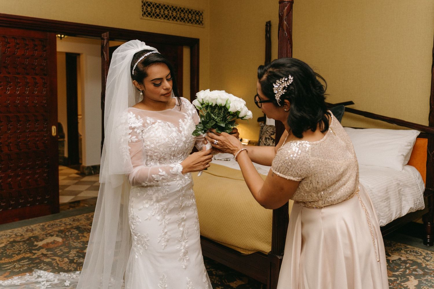 A bride gets final adjustments to her white lace wedding gown in a dimly-lit room before the ceremony.