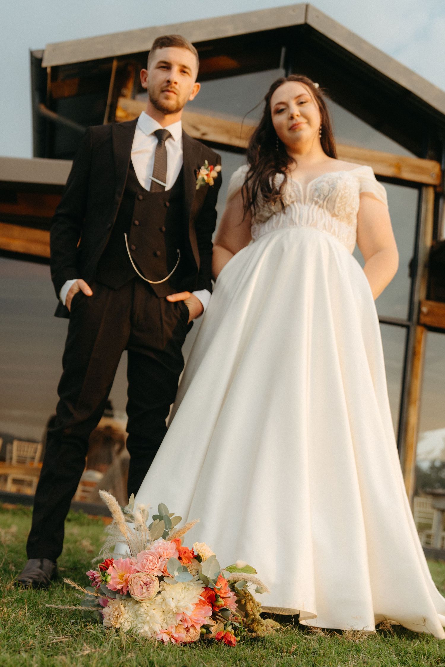 A couple poses together outdoors with a beautiful floral bouquet on the grass.