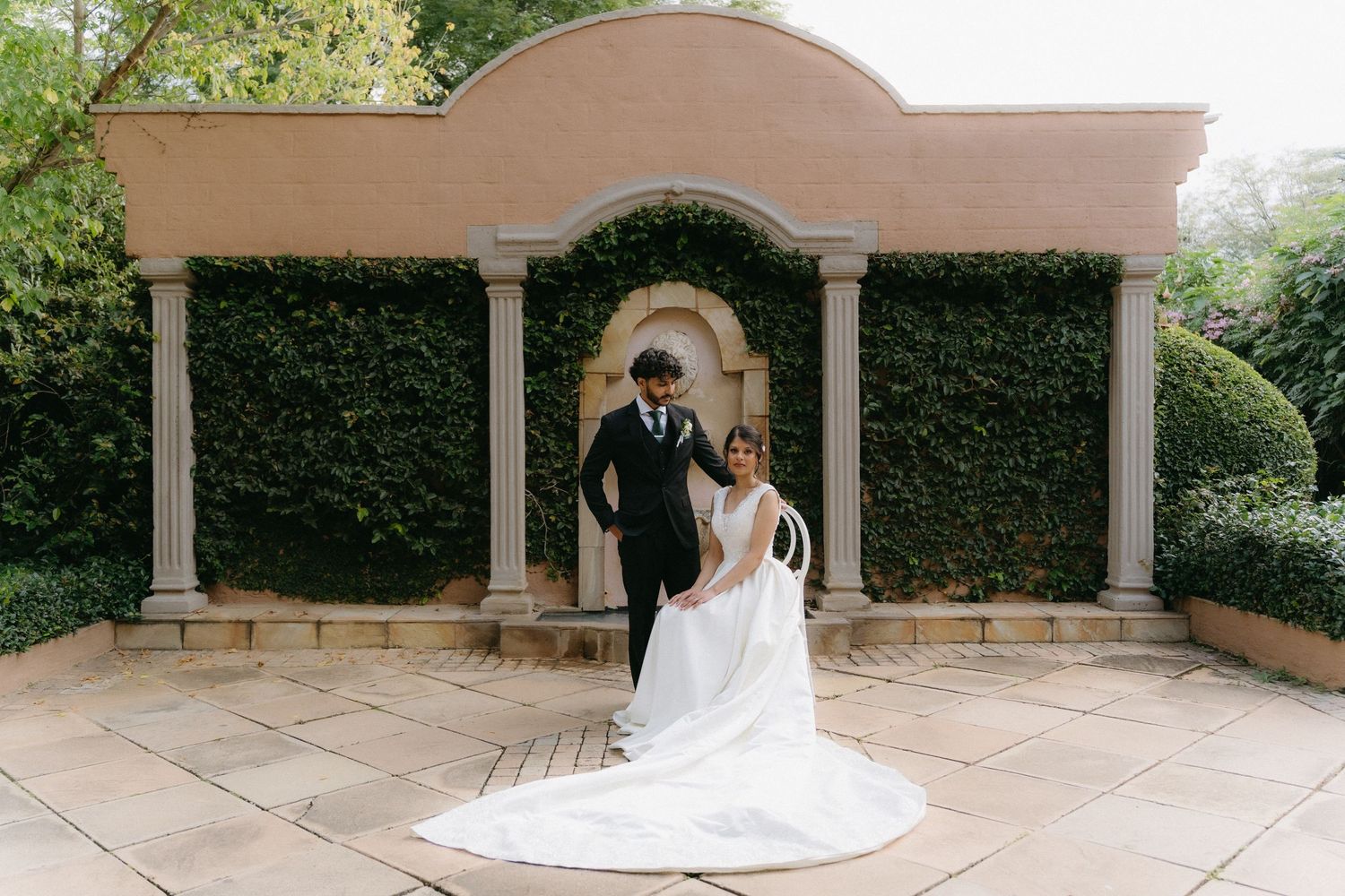 A couple in wedding attire pose under an ivy-covered stone archway in an elegant garden setting.