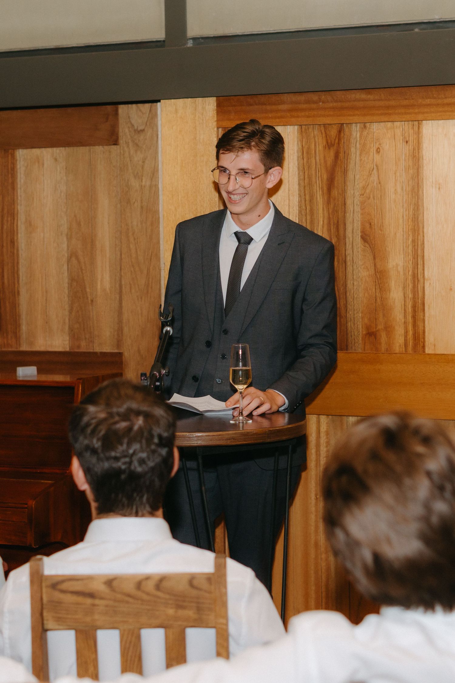 A speaker in a grey suit delivers a speech at a wooden podium during a wedding reception.