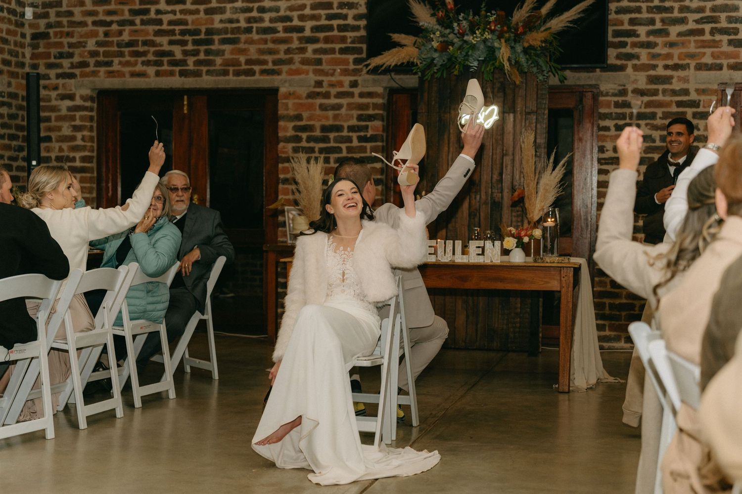 Wedding guests dance and celebrate at a rustic brick-walled reception venue with white chairs and warm lighting.
